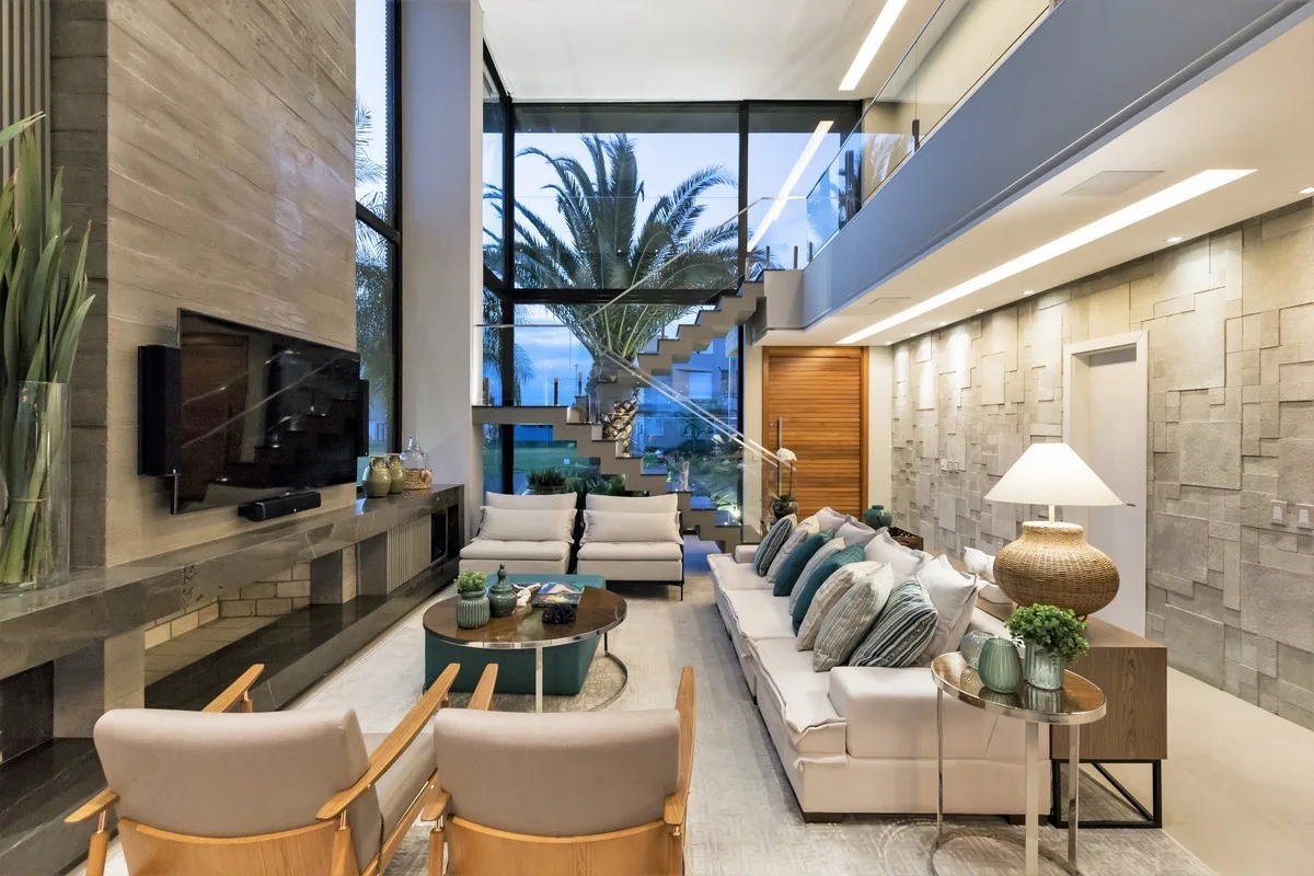 A spacious double-height living room featuring a long white sofa, a stone accent wall, a fireplace, and a floating glass staircase.