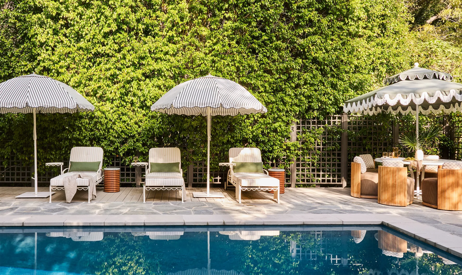 Three striped chaise lounges with green bolster pillows sit beside a swimming pool under scalloped umbrellas, backed by a tall green hedge.