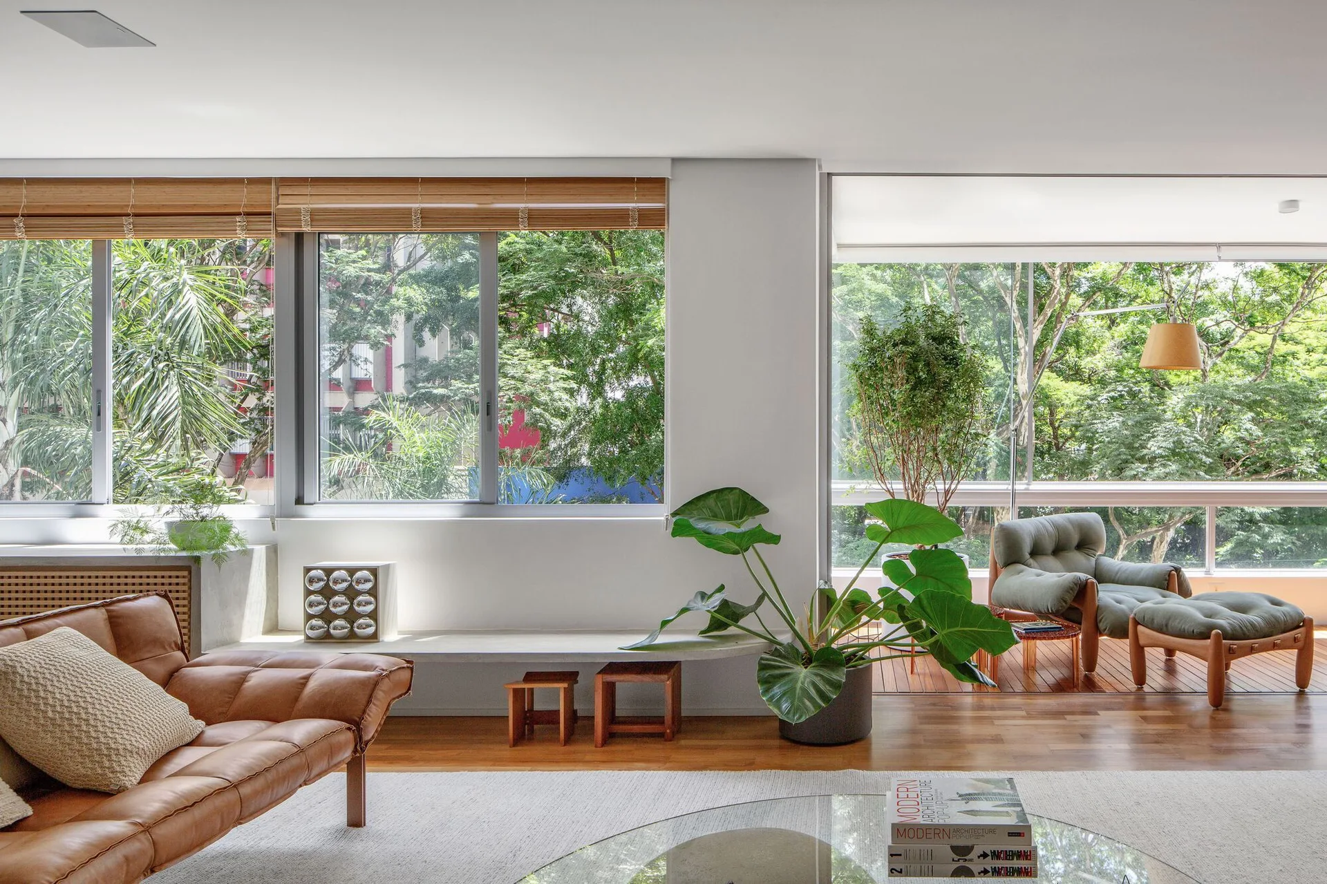 A sunlit living room features a tan leather sofa, a low concrete bench beneath large windows, and an adjacent balcony with an armchair.