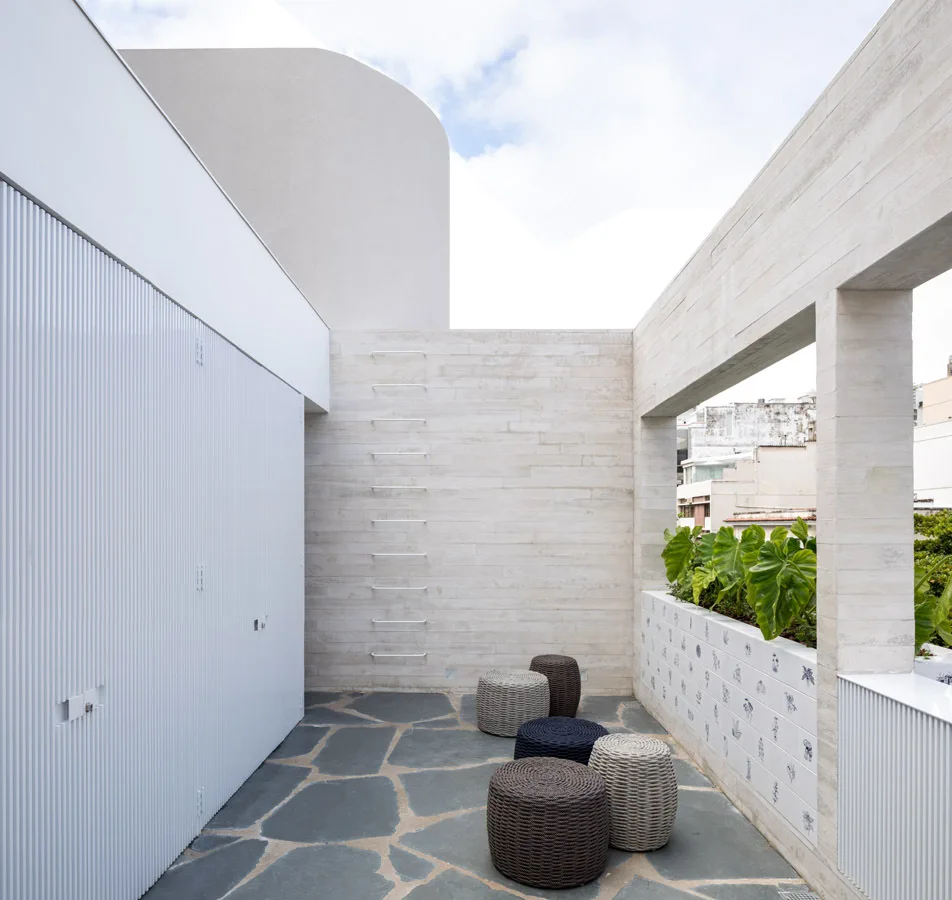 An outdoor terrace featuring irregular flagstone flooring, a white ribbed wall, a tiled planter with green plants, and several woven stools.