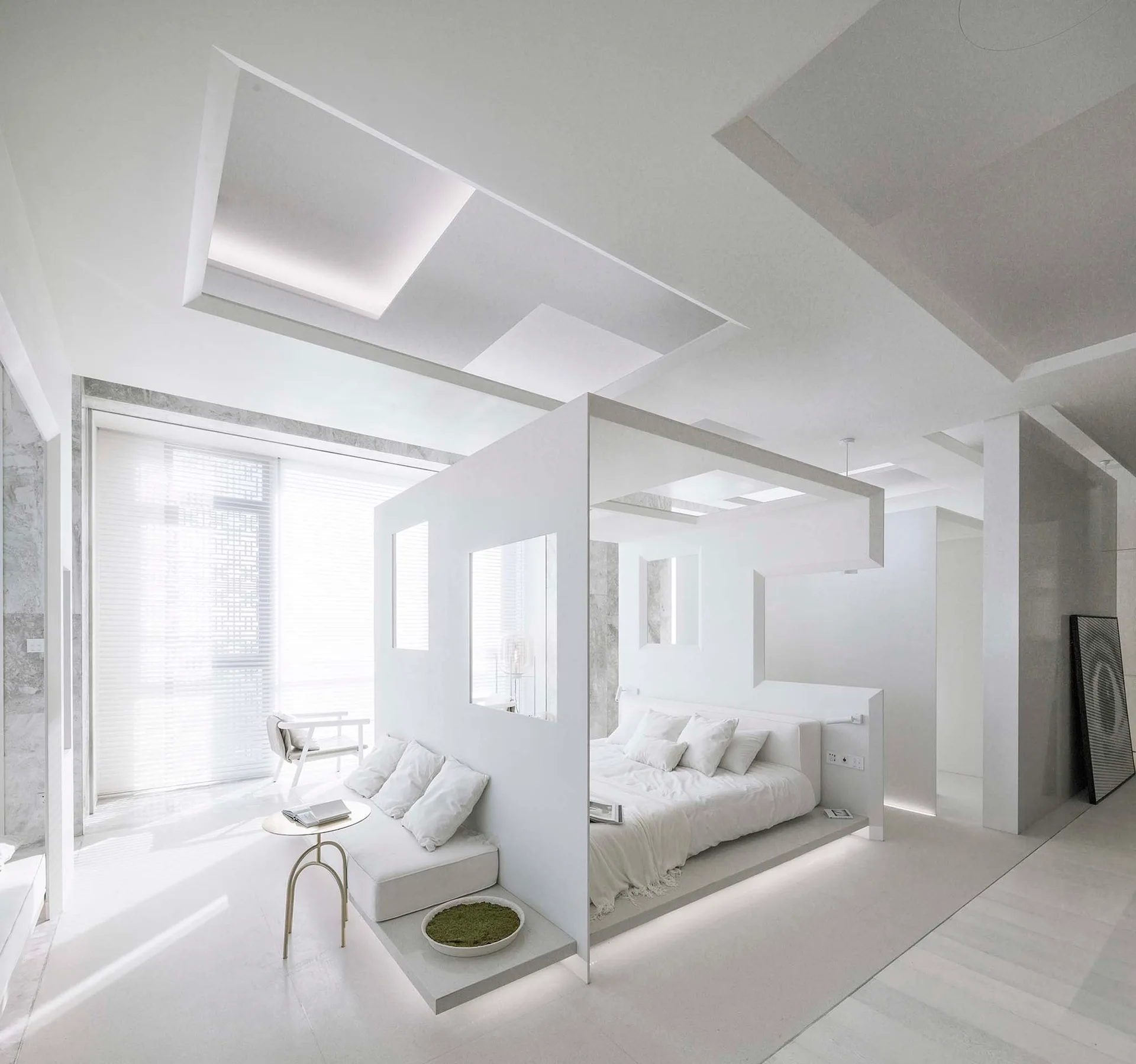 A minimalist white bedroom featuring a central geometric partition wall that separates a platform bed from a low-profile white sofa and side table.