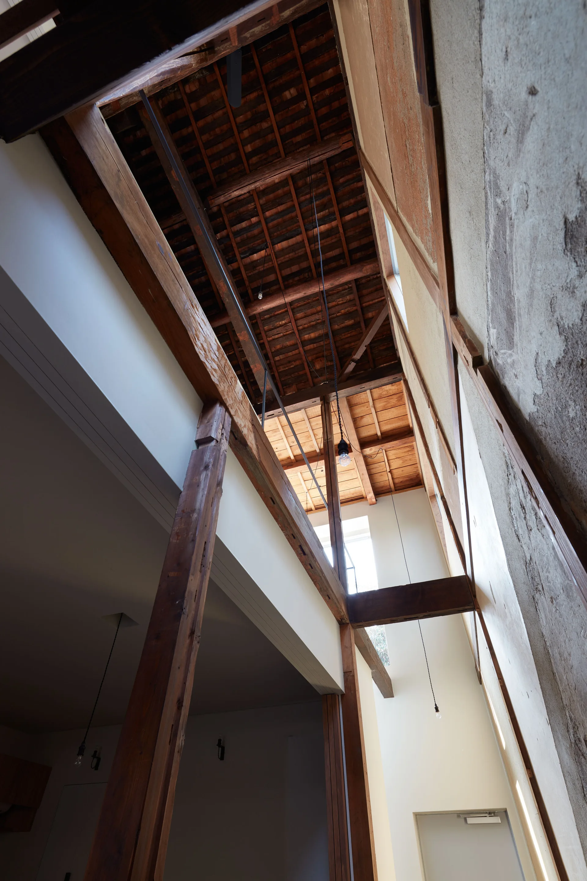 A double-height interior space features exposed wood ceiling rafters, structural timber columns, and unfinished plaster walls illuminated by natural light.