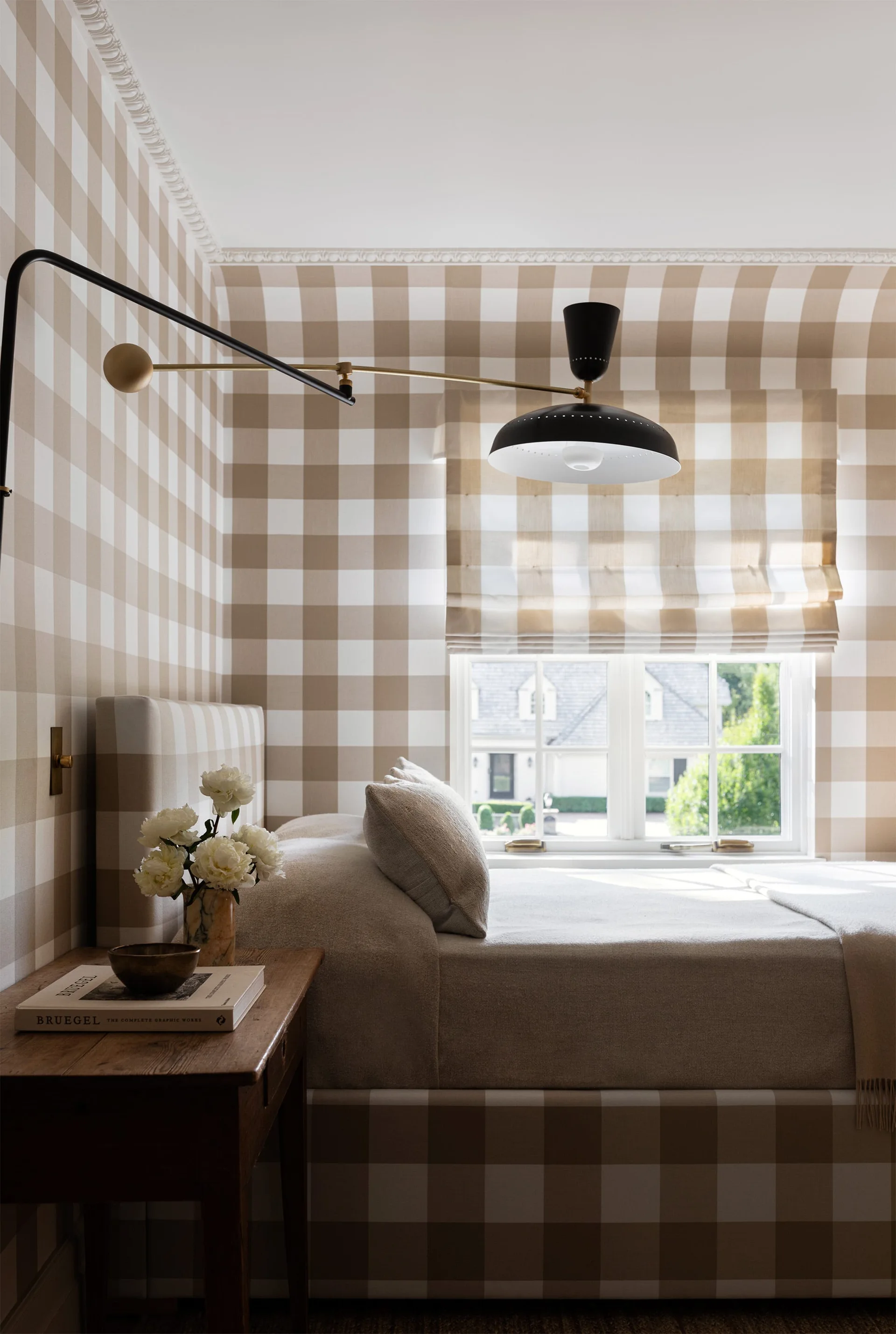 A bedroom featuring a bed, headboard, and walls upholstered in matching beige and white gingham fabric with a black and brass swing-arm wall lamp.