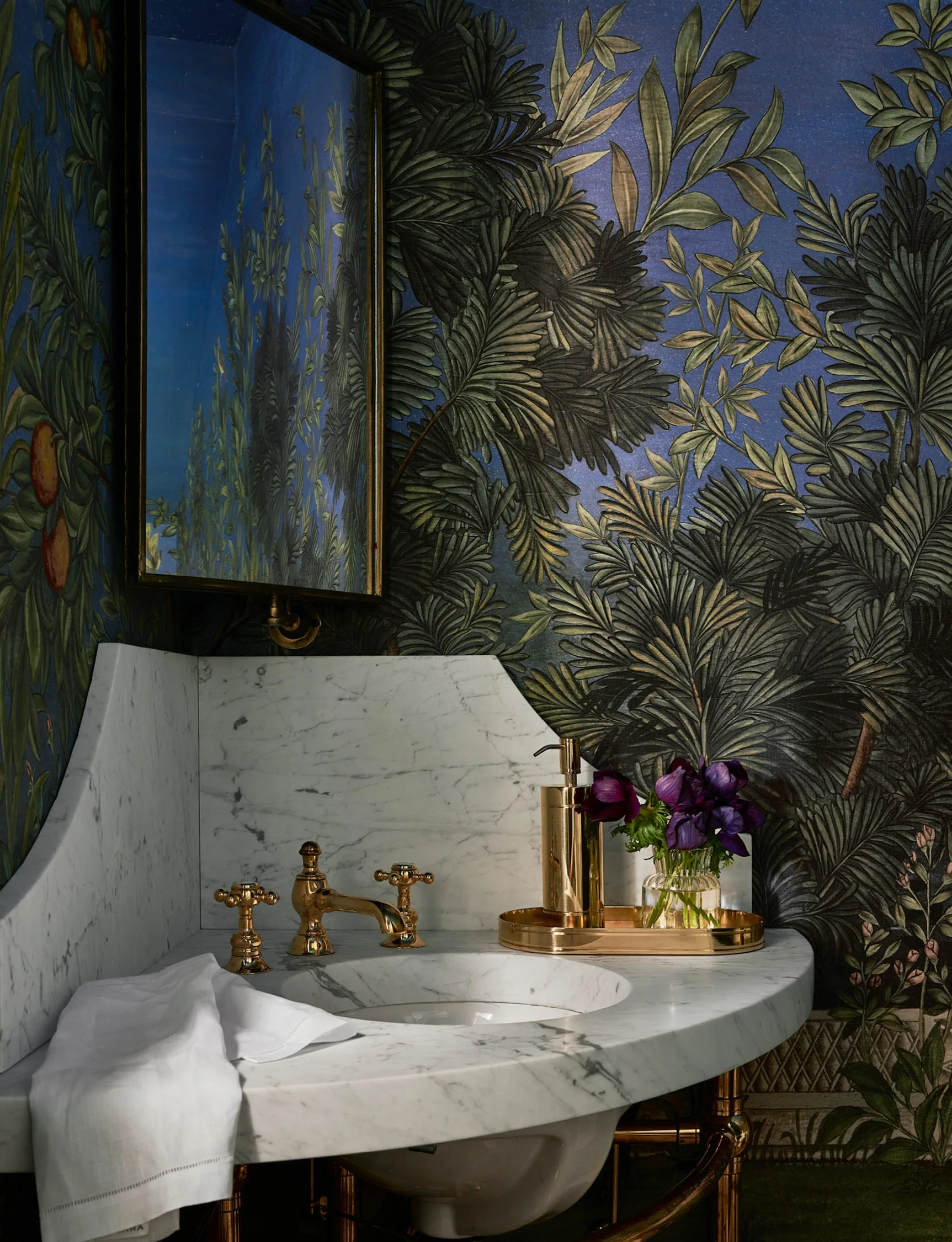 A white marble corner washstand with brass fixtures set against dark green and blue botanical wallpaper in a moody powder room.
