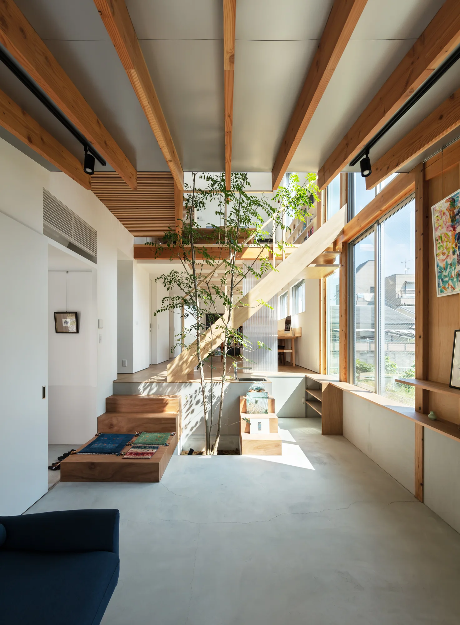 A modern split-level living space features a central green tree growing from the concrete floor, surrounded by wooden stairs, beams, and large windows.