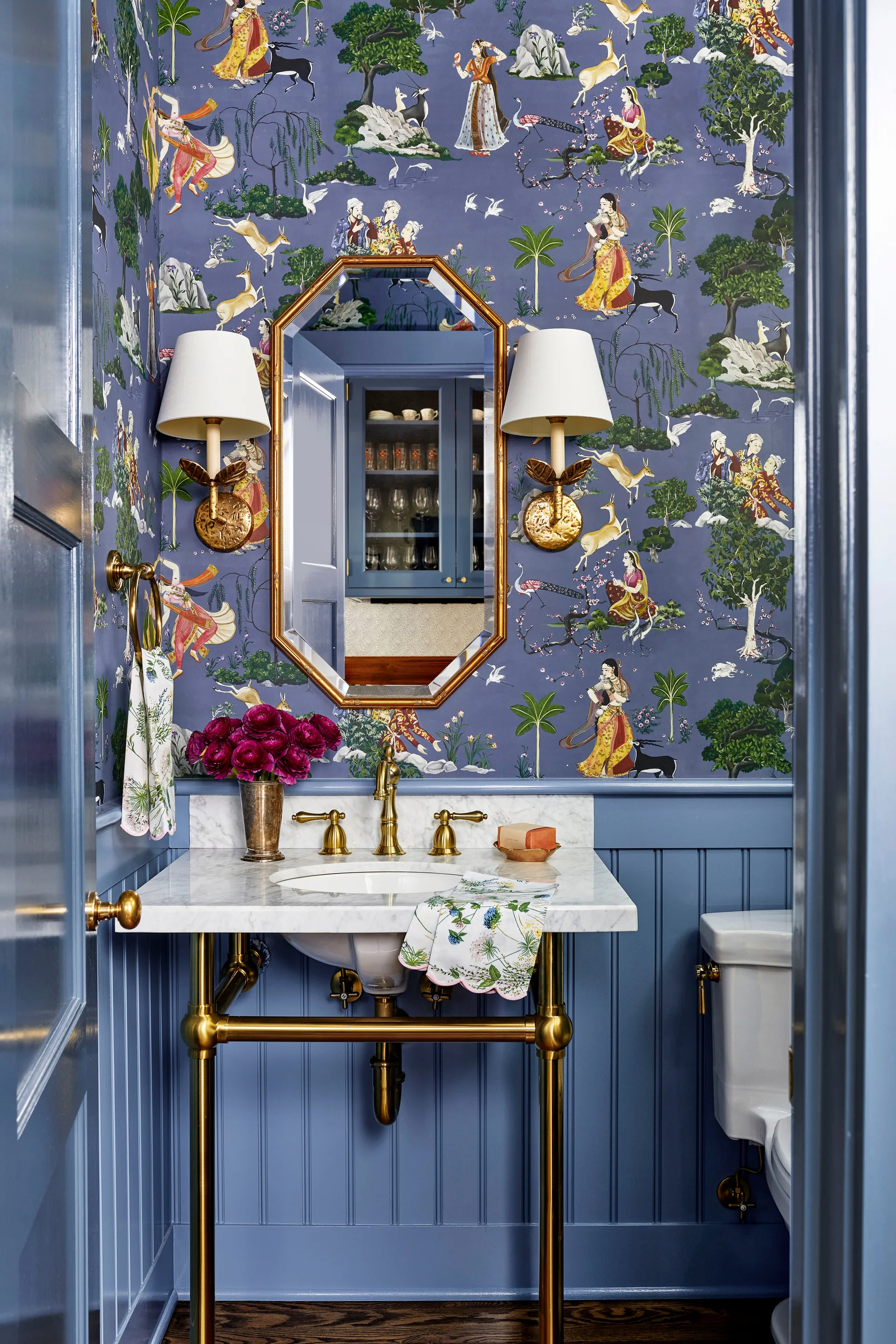 A blue powder room features pictorial wallpaper above matching beadboard wainscoting, an octagonal gold mirror, and a brass console sink.
