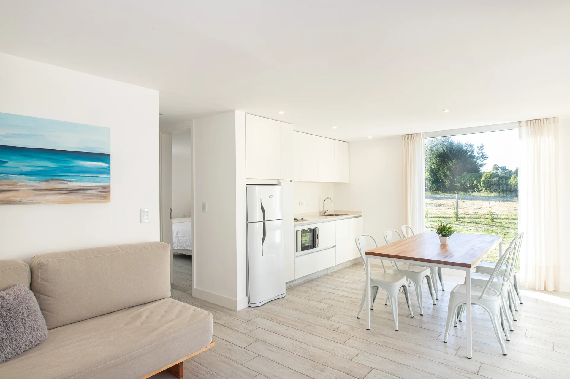 A bright open-plan space containing a white modern kitchen, a wooden dining table with metal chairs, and a window with sheer curtains.