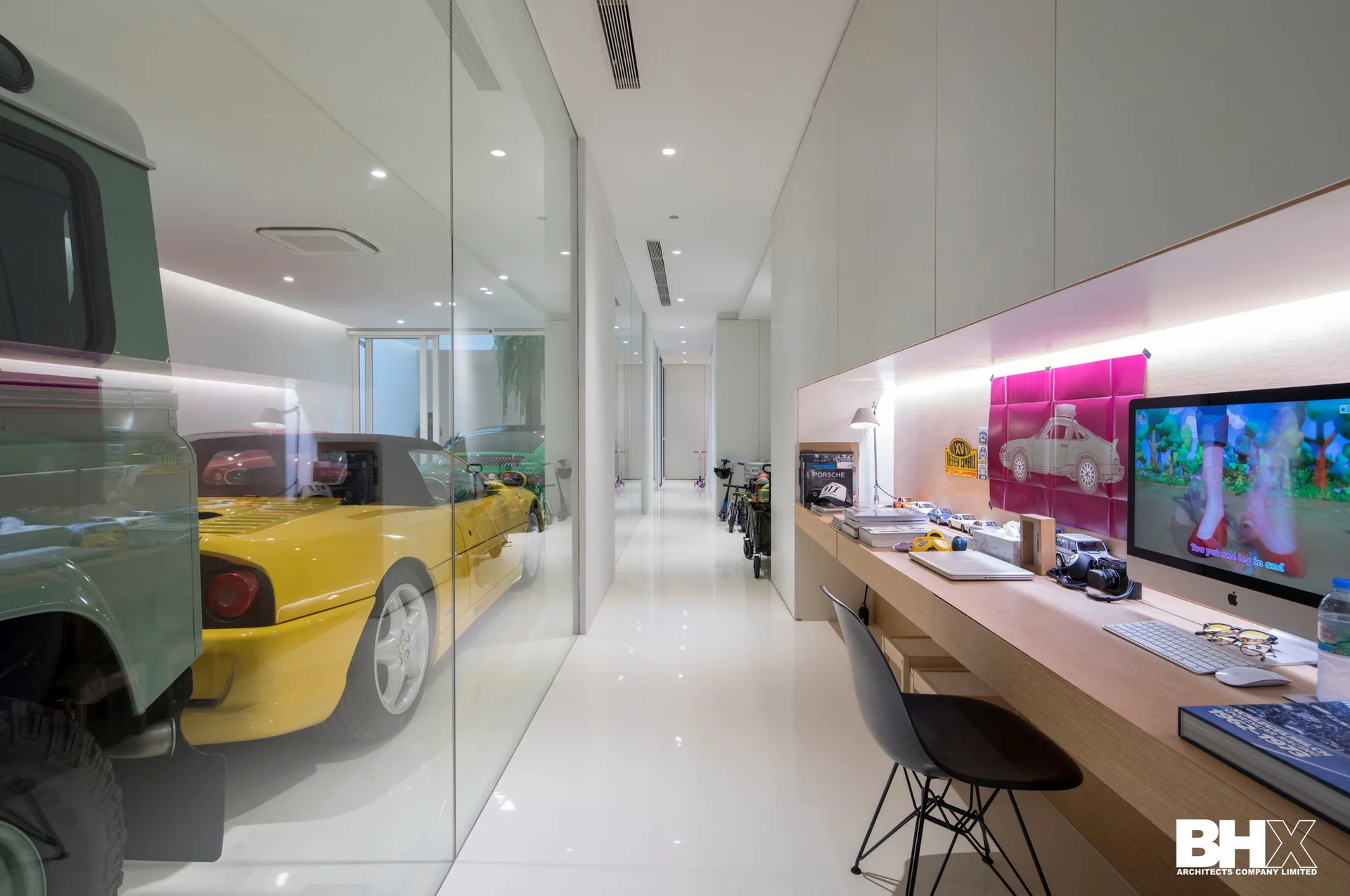 A modern home office hallway features a built-in wood desk under white cabinets and a full-height glass wall displaying cars in an adjacent garage.