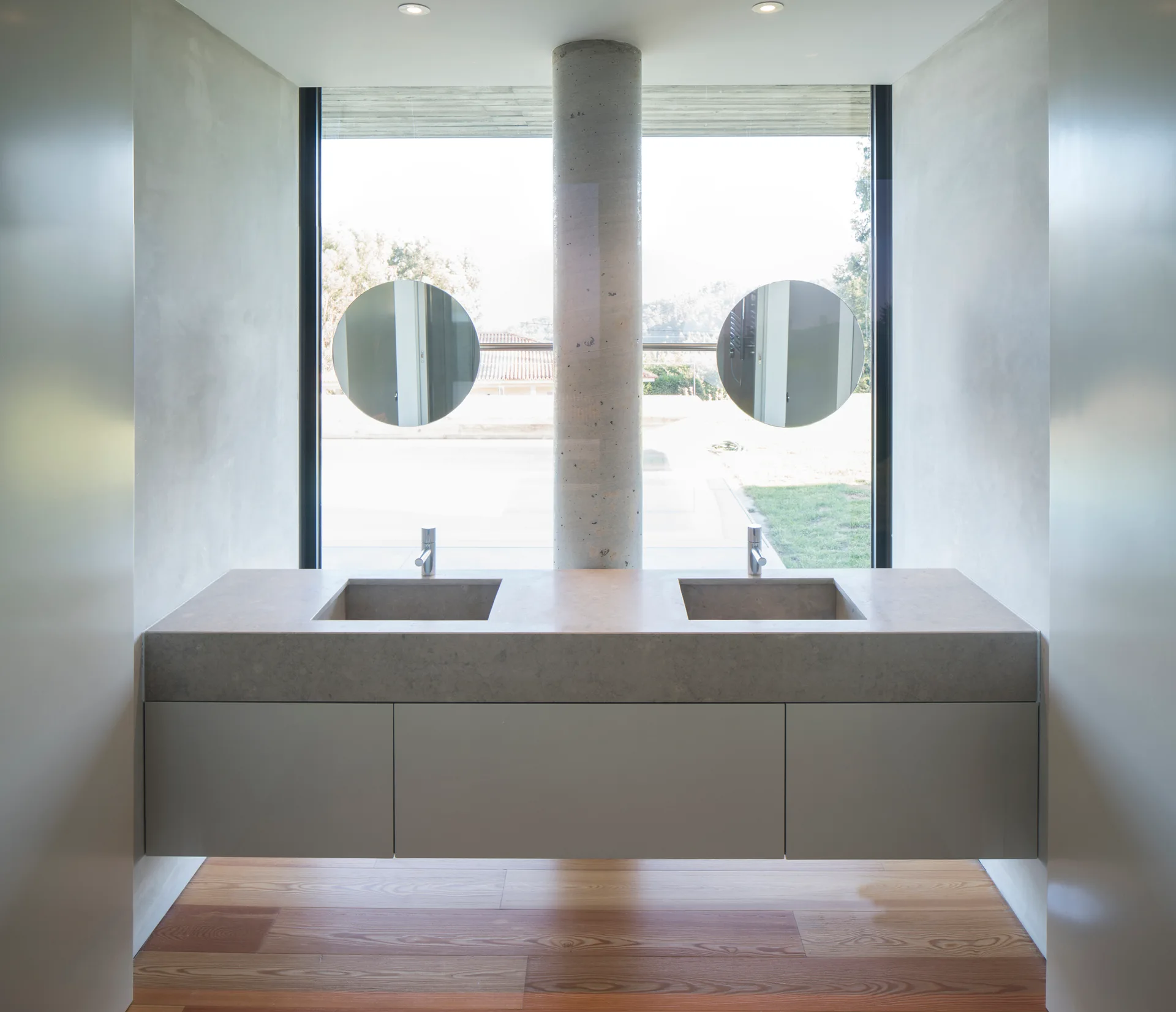 A minimalist bathroom features a floating double-sink vanity in front of a large window with a central concrete column and circular mirrors.