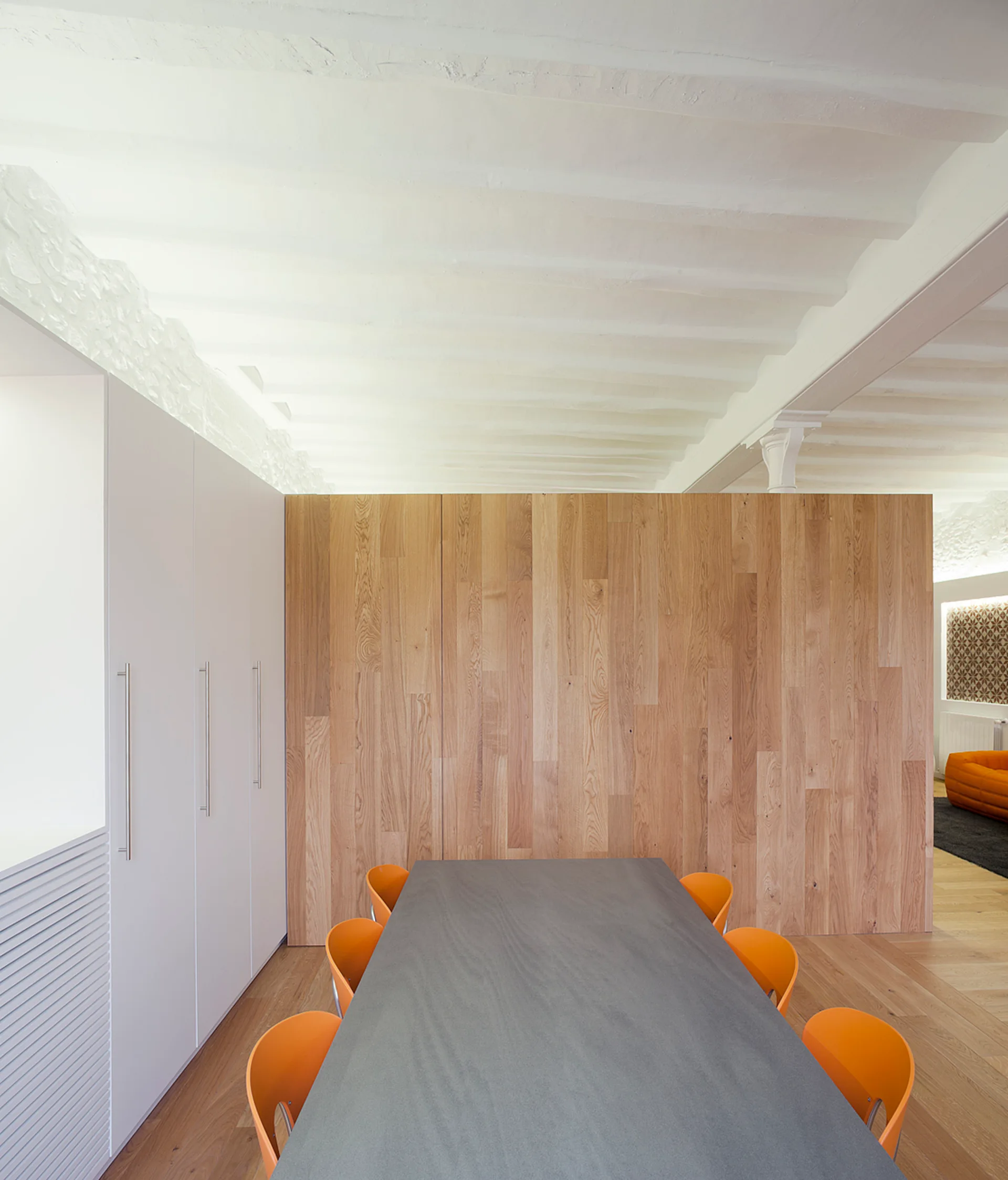 A grey dining table paired with bright orange chairs sits before a vertical wood-paneled partition wall under a white beamed ceiling.