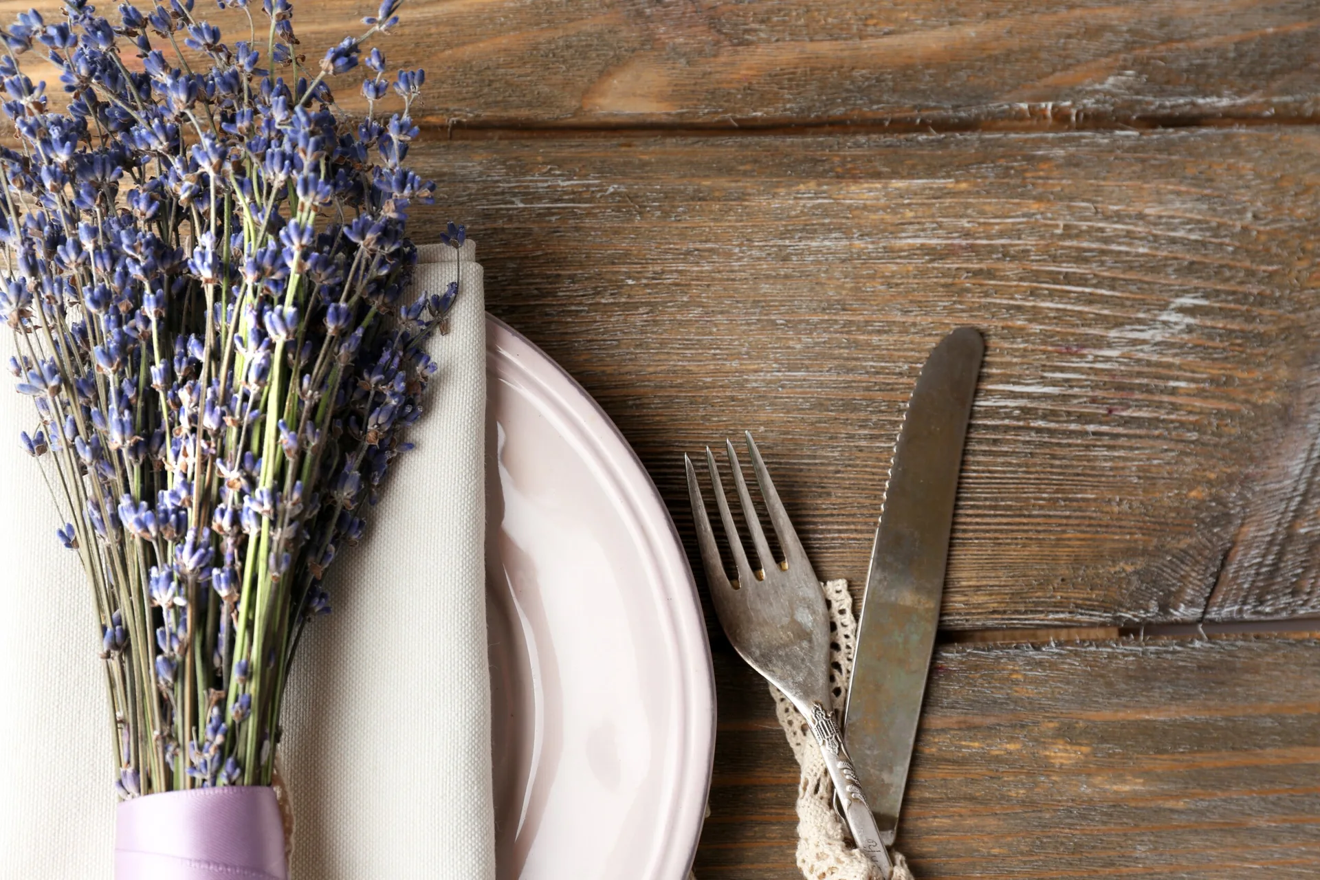 A rustic wooden tabletop decorated with a pale pink plate, vintage silverware, a linen napkin, and a bouquet of dried lavender.