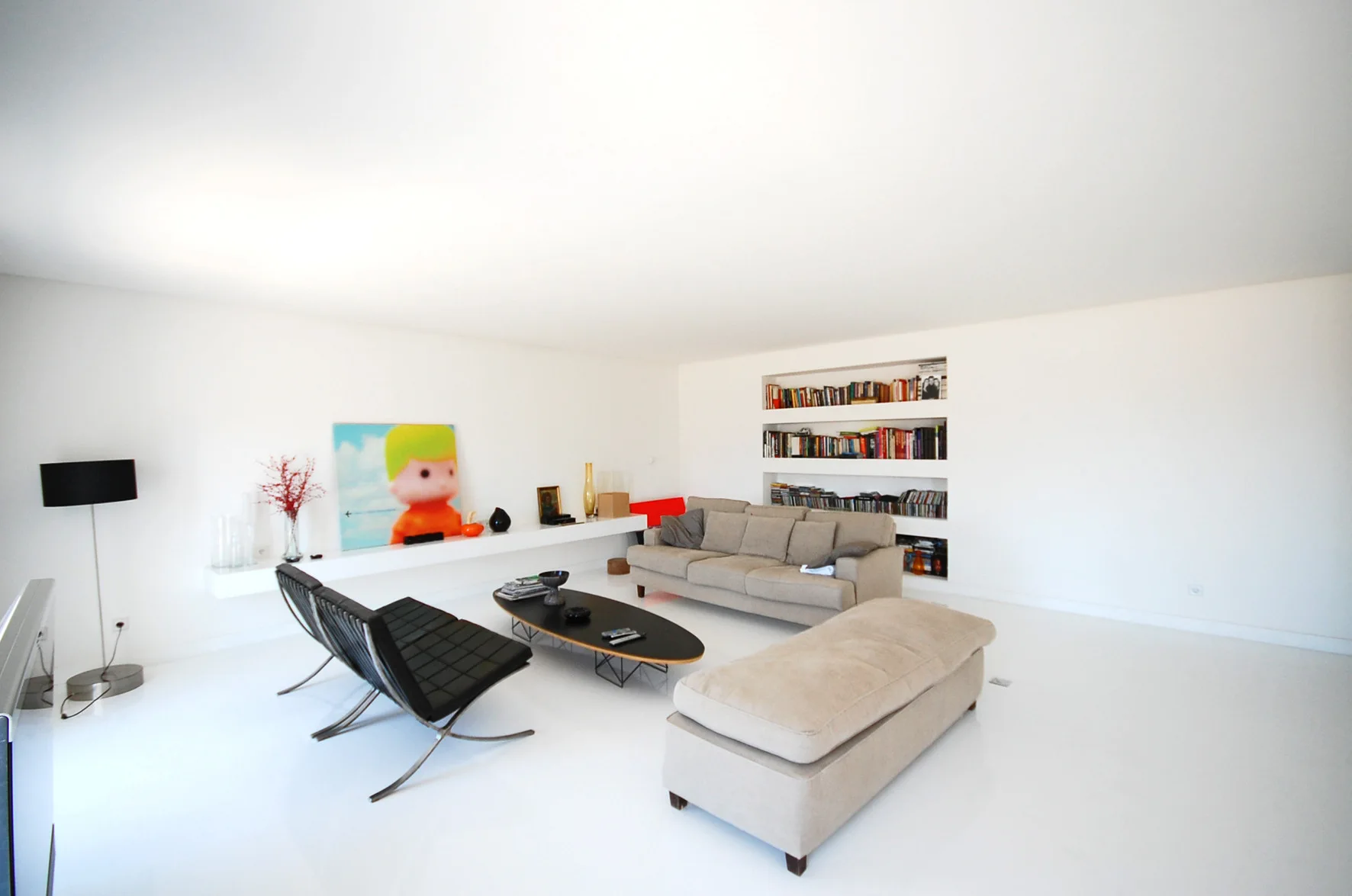 A minimalist living room with a glossy white floor, a beige sofa, black leather lounge chairs, and recessed wall shelving filled with books.