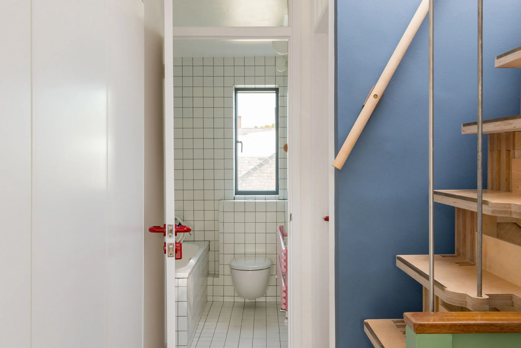 A view through an open white doorway with a red handle into a small bathroom with white grid tiles, next to a plywood staircase and blue wall.