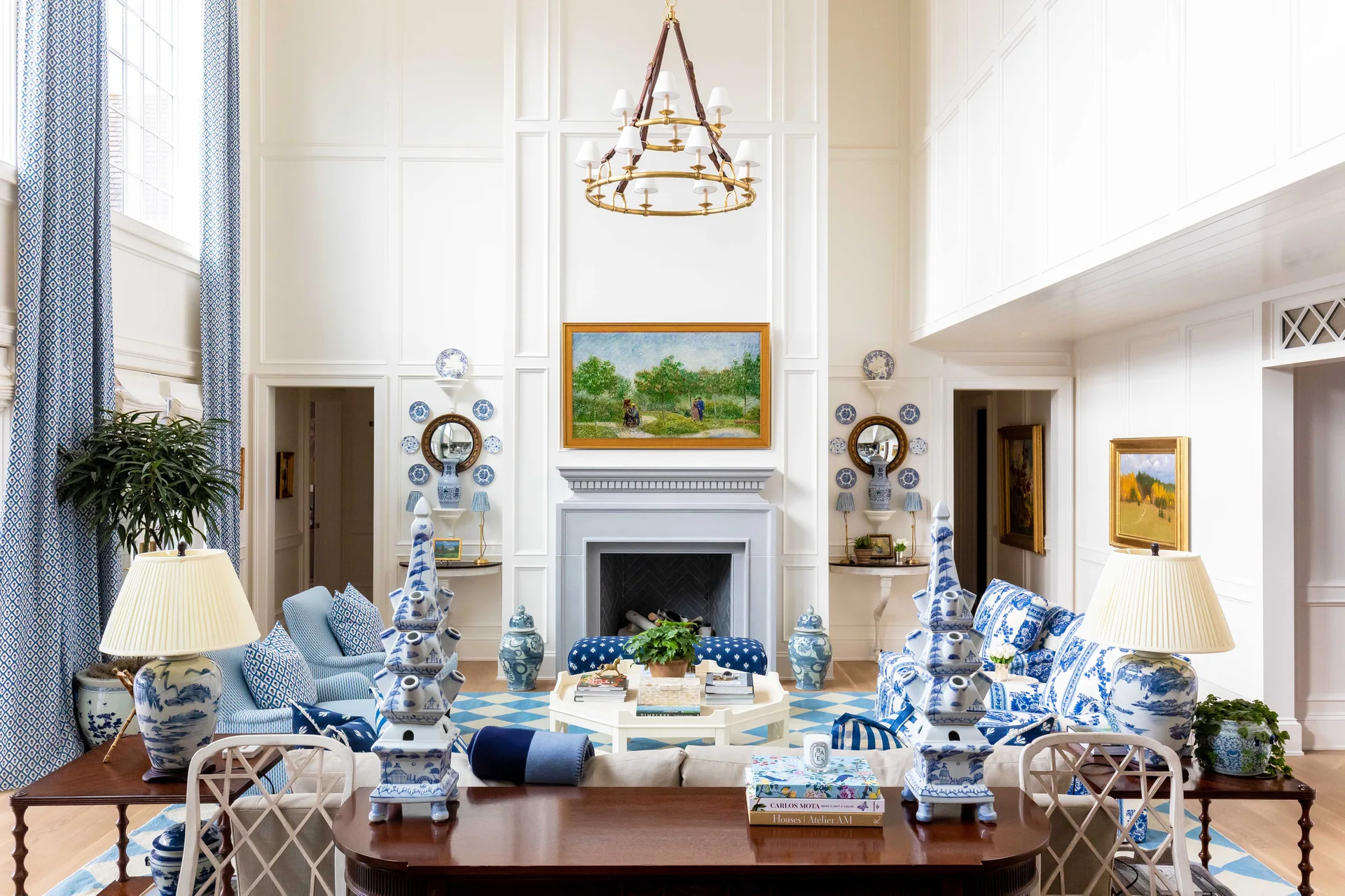 A high-ceilinged living room features white paneled walls, a central fireplace, and symmetrical blue and white patterned furniture and decor.