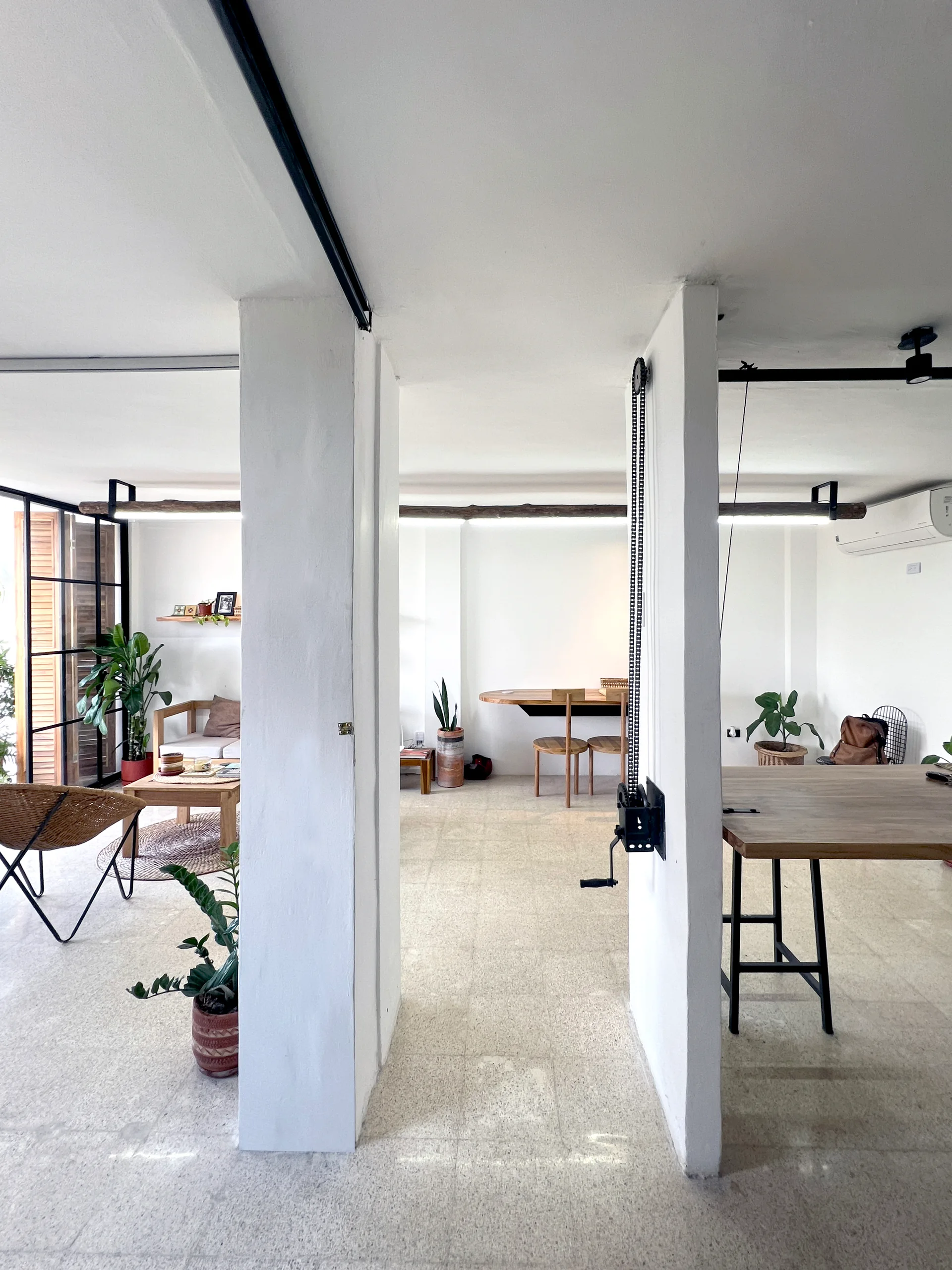 White structural pillars divide an open-plan living room featuring terrazzo floors, wooden furniture, and potted houseplants.