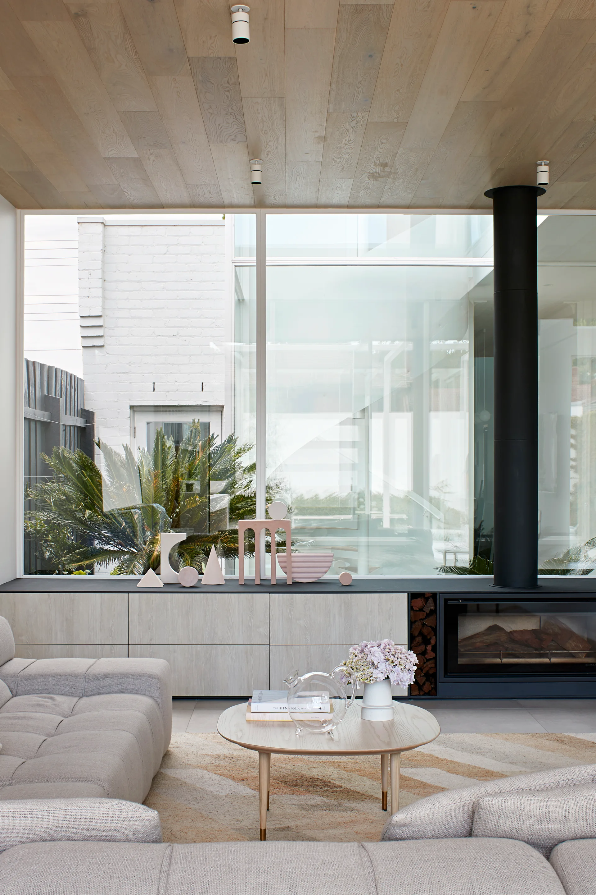 A modern living room features a grey modular sofa, a light wood coffee table, a wooden plank ceiling, and a low storage cabinet with an integrated fireplace.