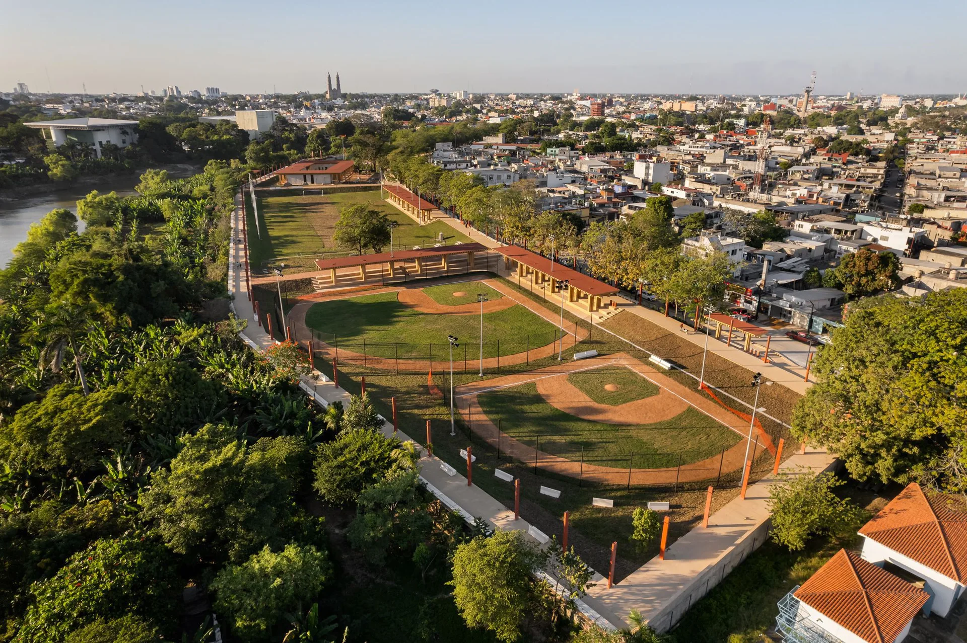 An aerial view of an urban sports park featuring baseball fields, covered walkways with terracotta roofs, concrete pathways, and surrounding trees.