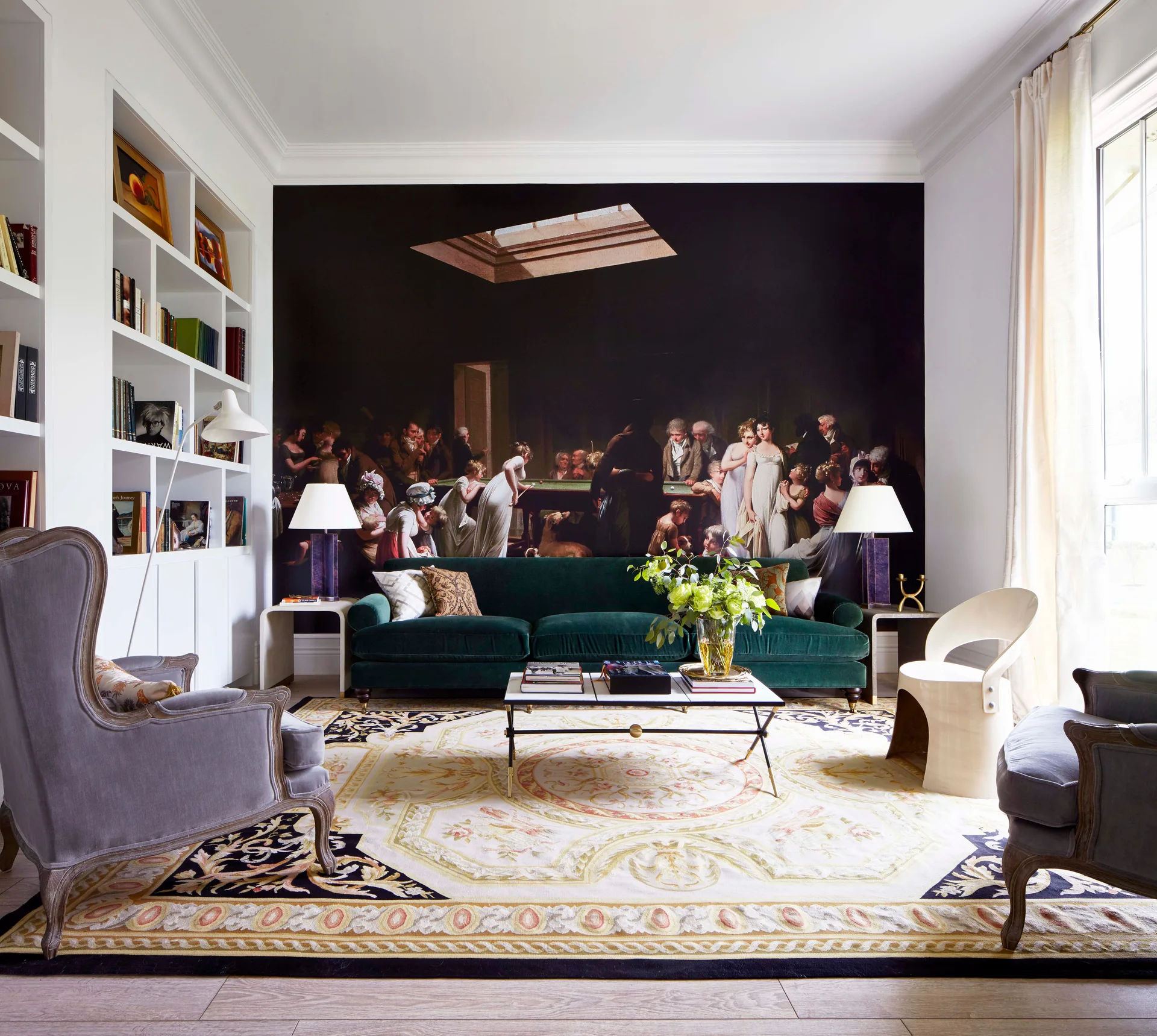 A living room features a deep green velvet sofa set against a large classical wall mural, flanked by white bookshelves and an ornate patterned rug.