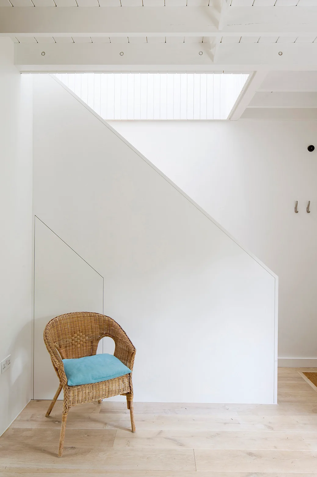 A minimalist white hallway features a wicker armchair with a blue cushion beneath an angled staircase wall with integrated under-stairs storage.