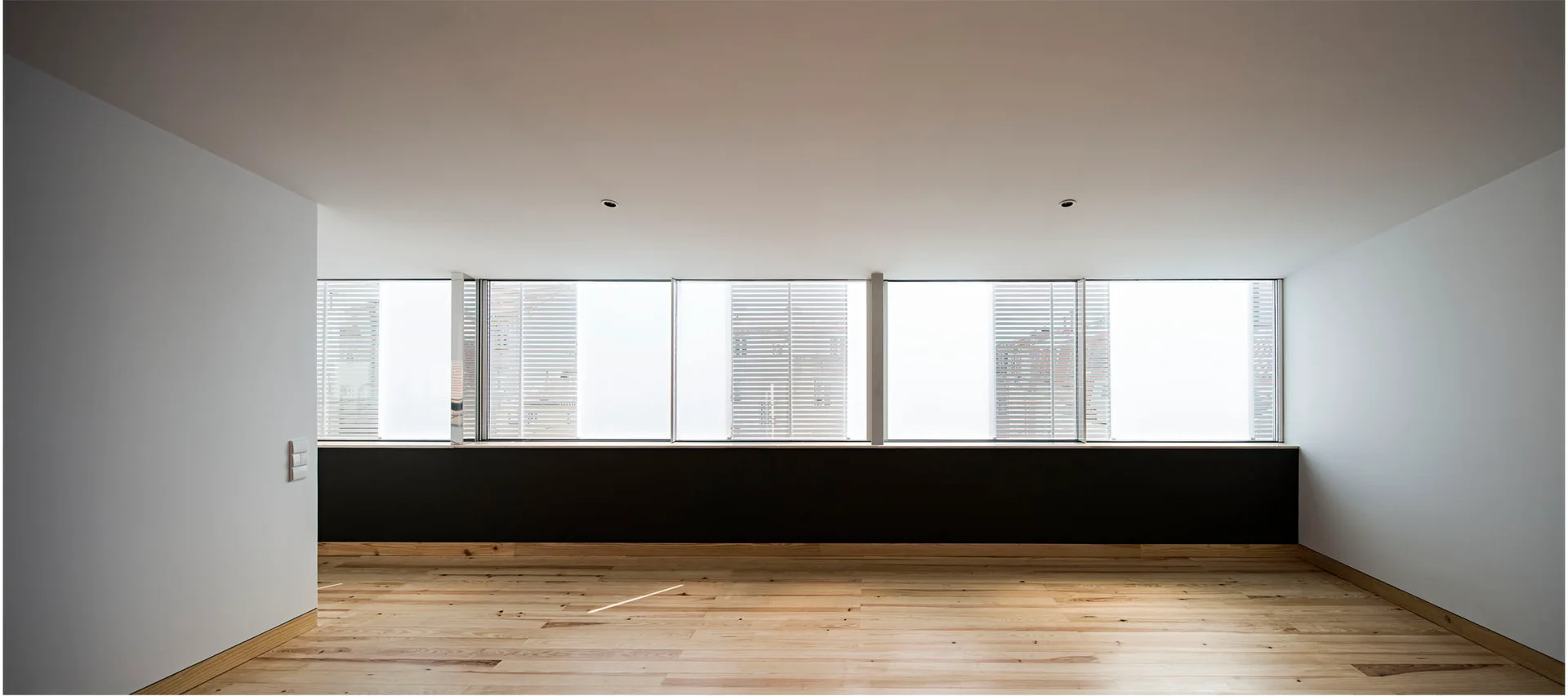 An empty minimalist room featuring a long horizontal window with a dark sill, white walls, and light-toned wood plank flooring.