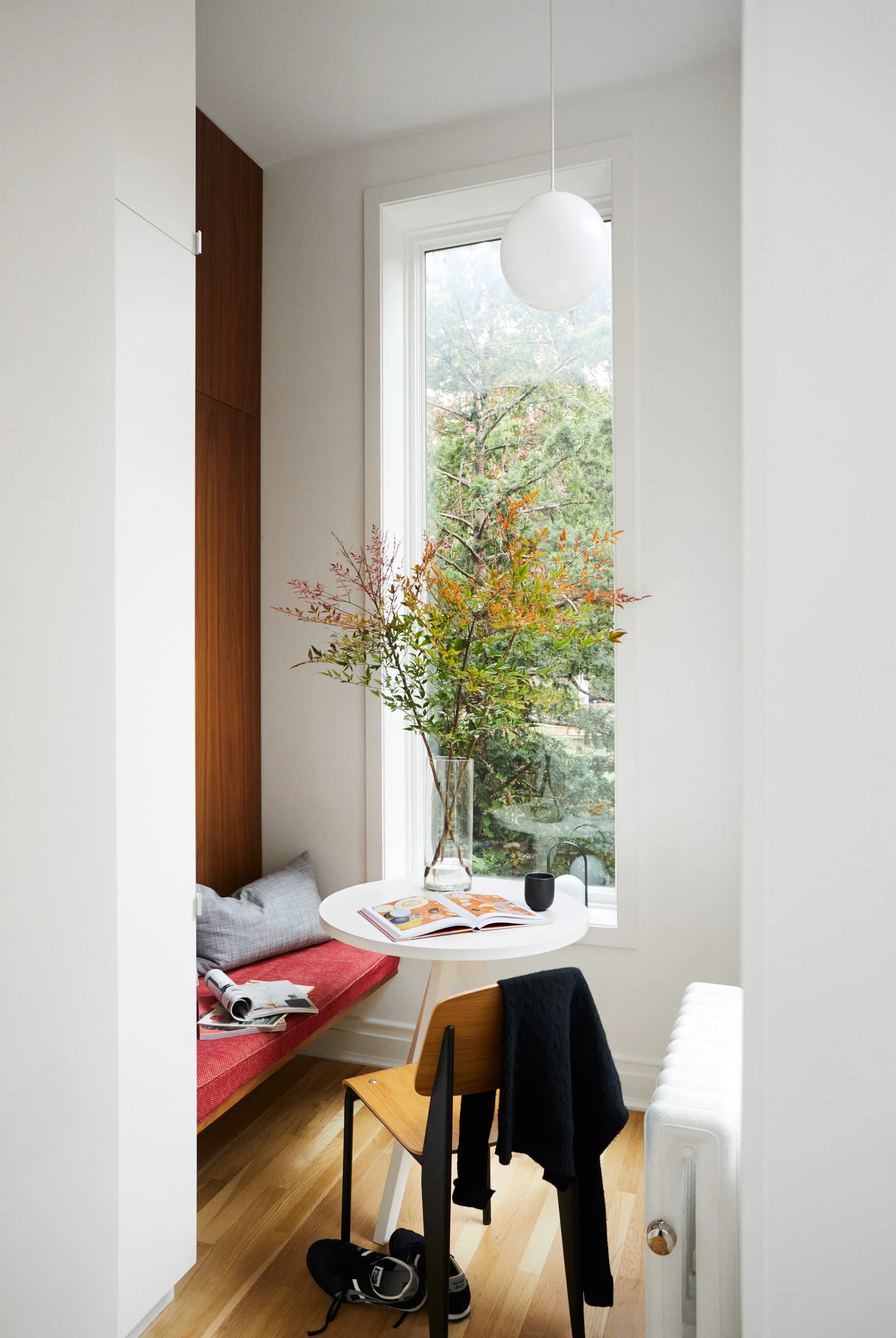 A small dining nook features a built-in bench with a red cushion, a small round white table, a wooden chair, and a tall window.