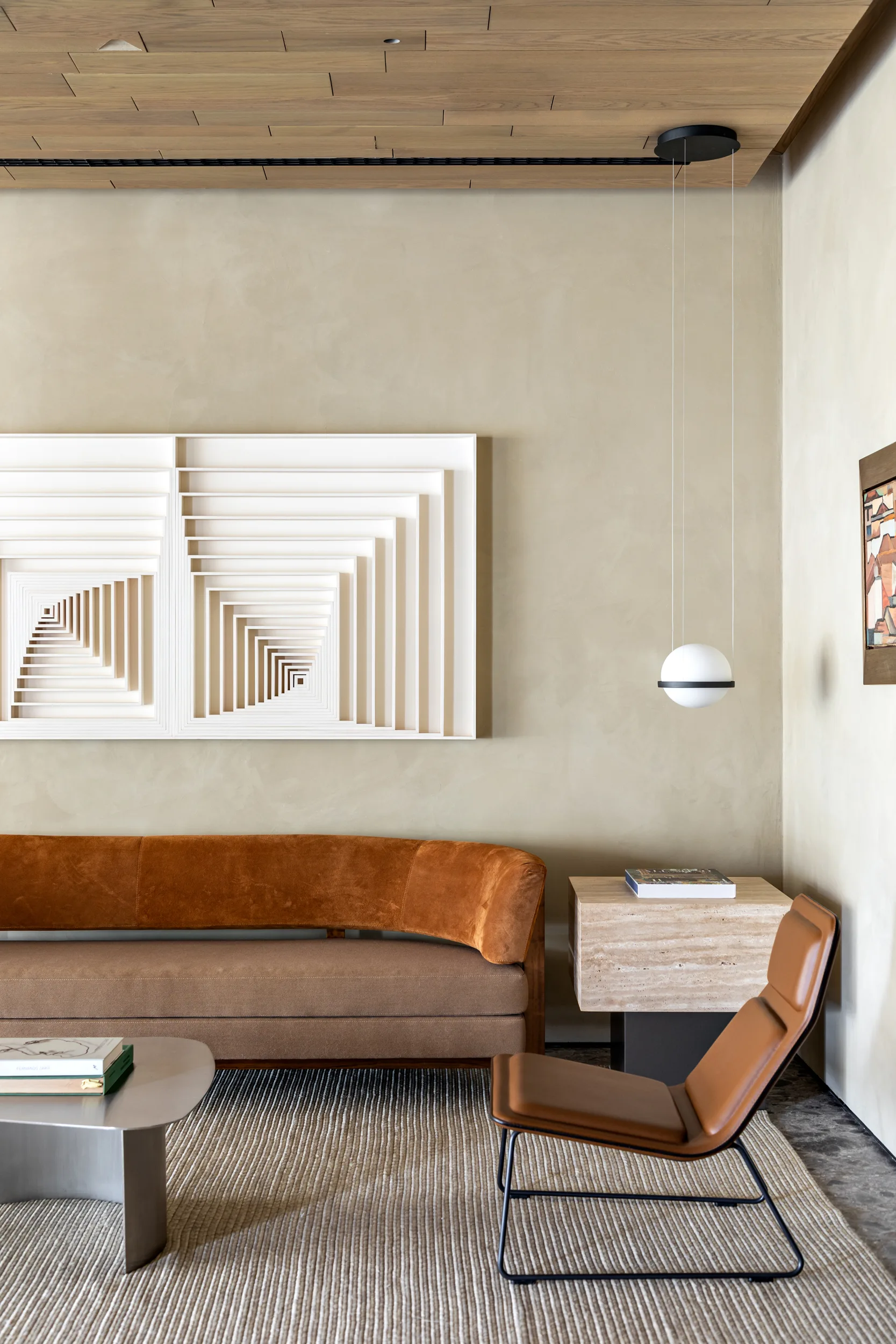 A contemporary living room features a tan sofa, leather accent chair, travertine side table, and a large white geometric wall sculpture.