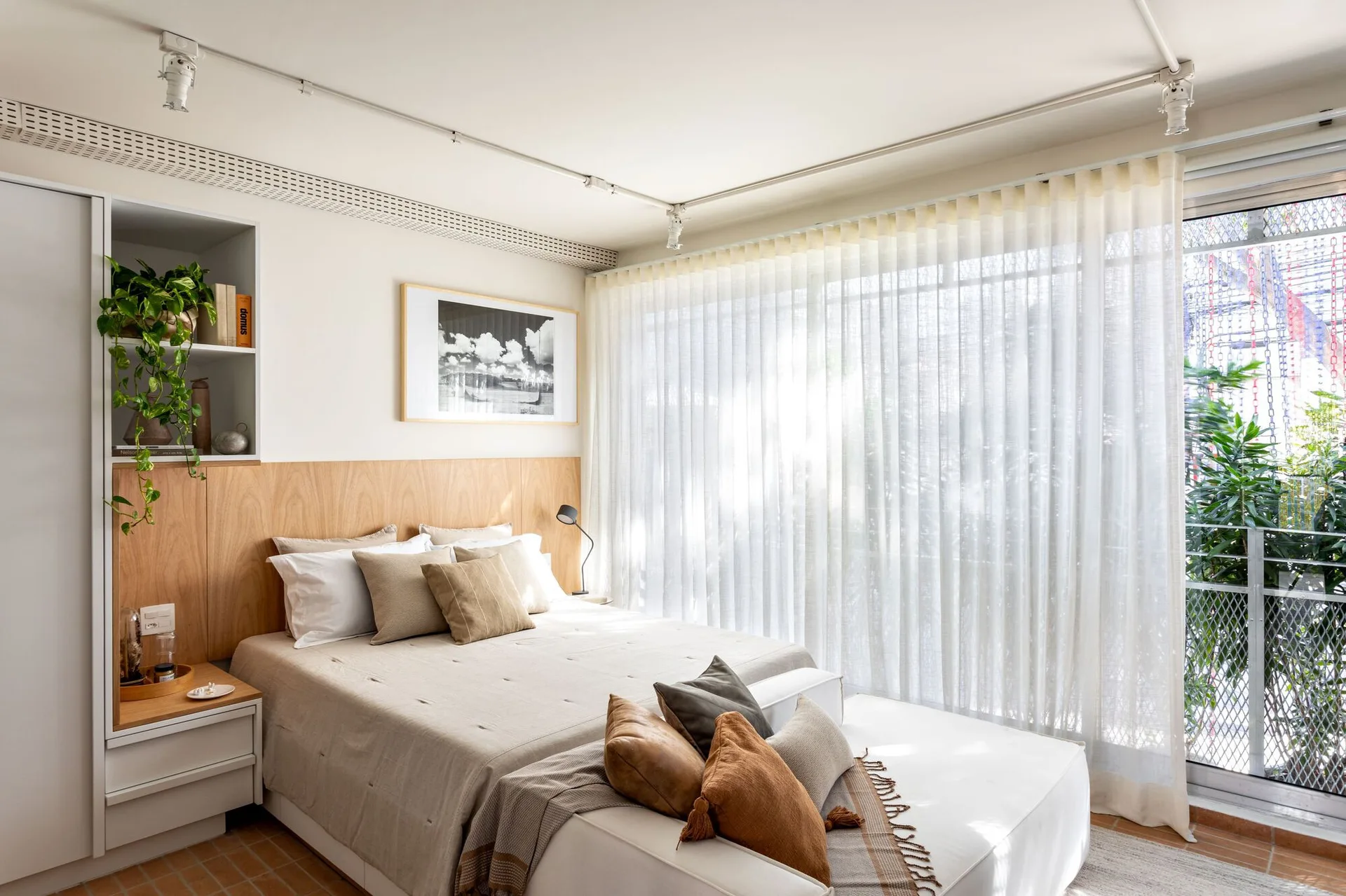 A modern bedroom features a low bed with a wood panel headboard, built-in shelving with a trailing plant, and sheer white curtains over a large window.
