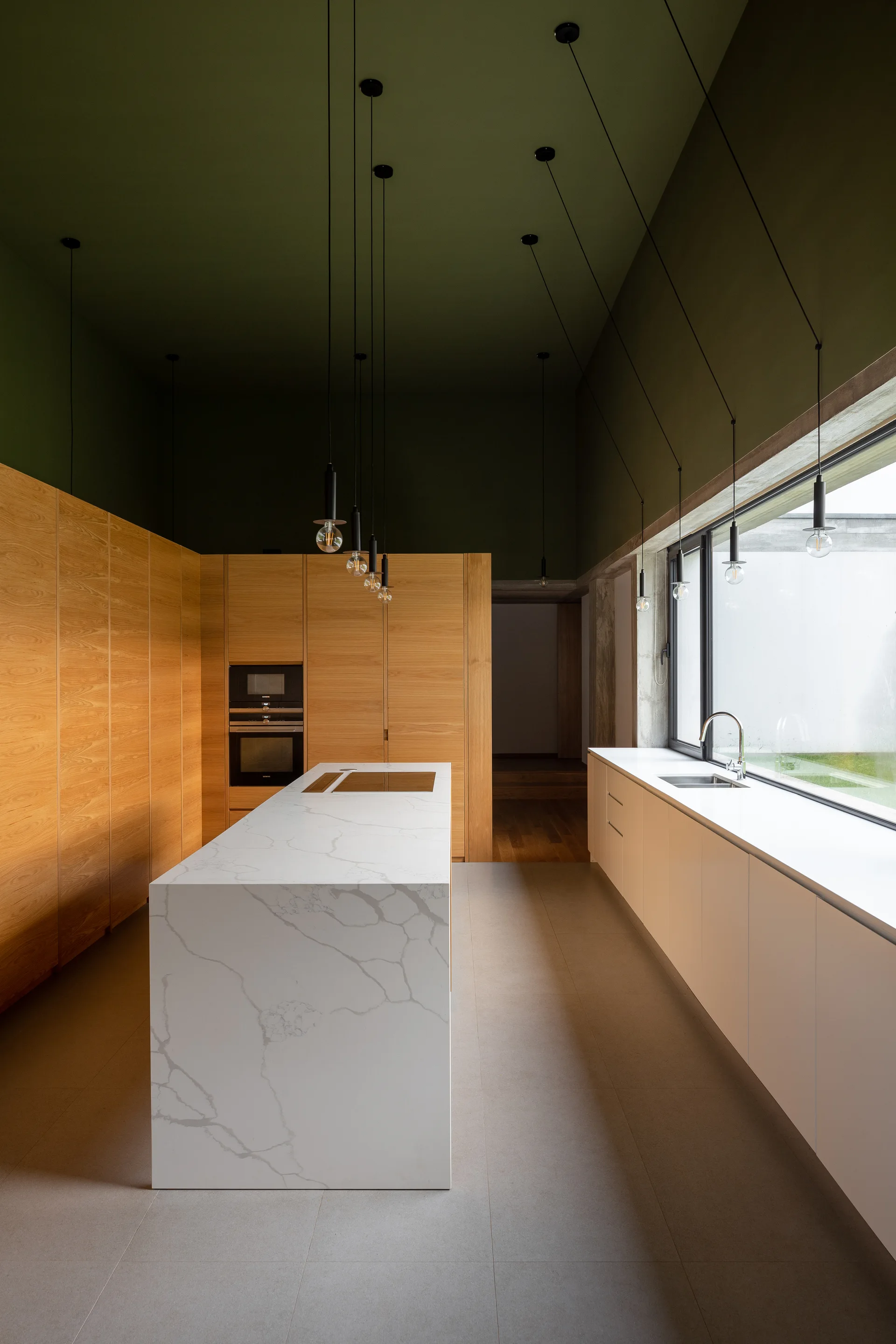 A modern kitchen featuring a white veined marble island, tall wood cabinetry, a dark green painted ceiling, and minimalist hanging pendant lights.
