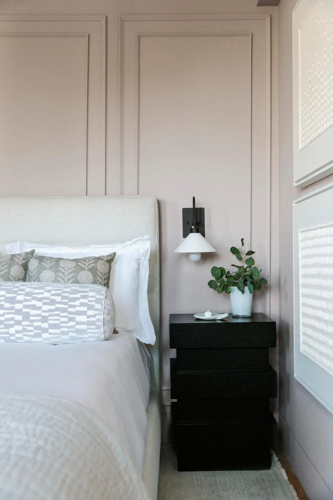 A bed with a light headboard sits beside a blocky black nightstand against a dusty mauve wall with classic picture frame molding.