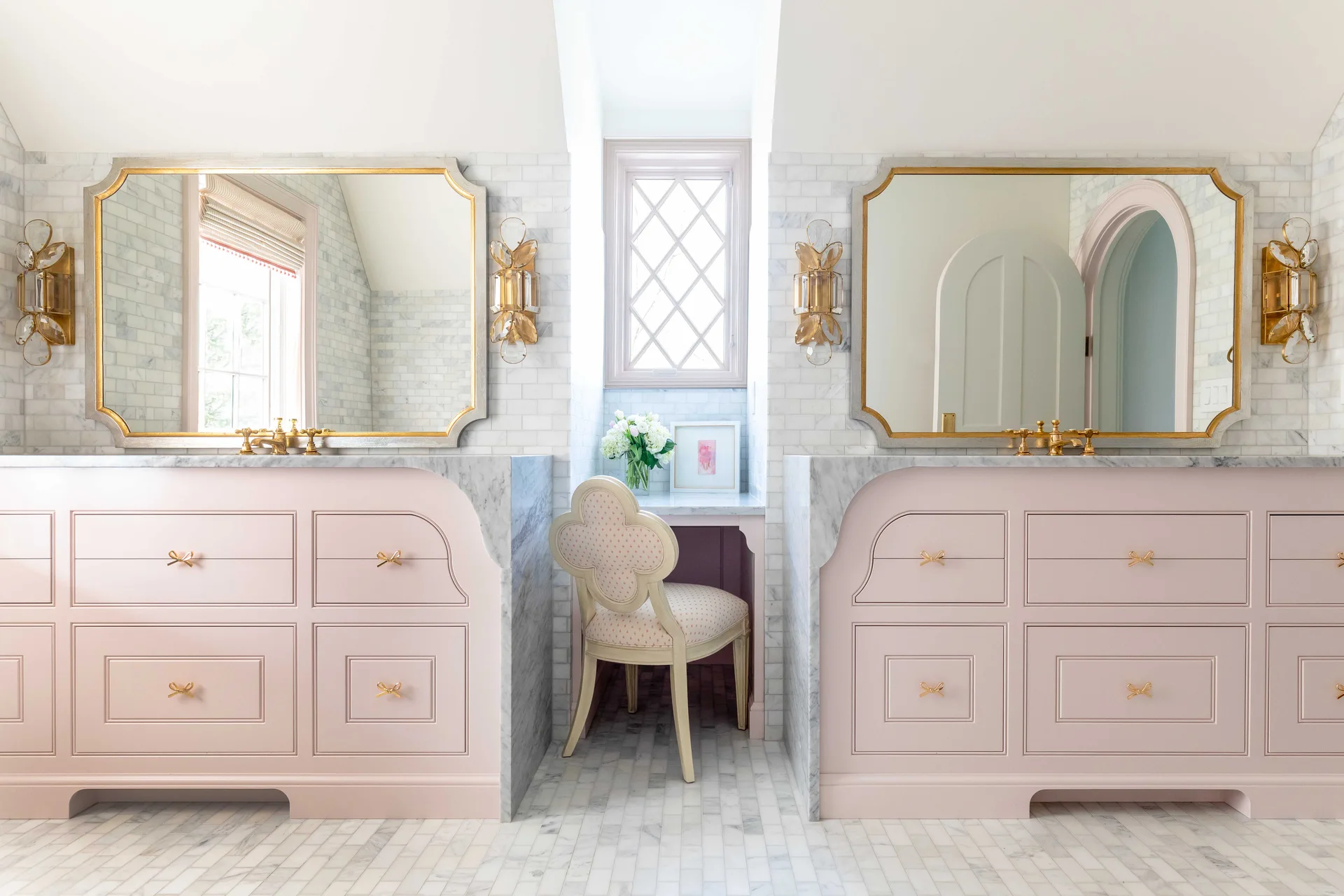 Two pastel pink vanities with marble countertops flank a central makeup desk with a patterned chair in a marble-tiled bathroom.