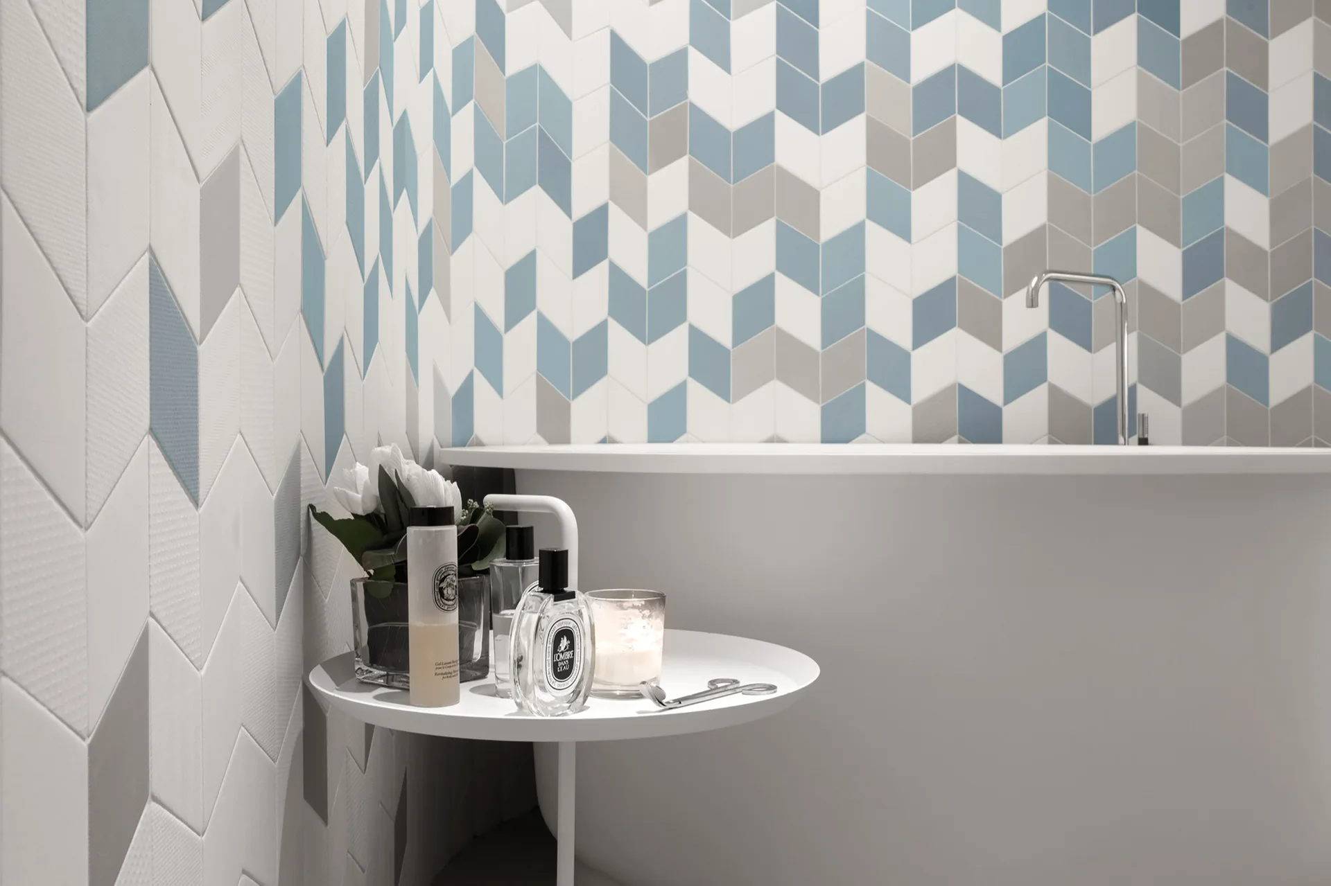 A white freestanding bathtub stands against a blue and grey chevron tile wall next to a small side table holding toiletries.