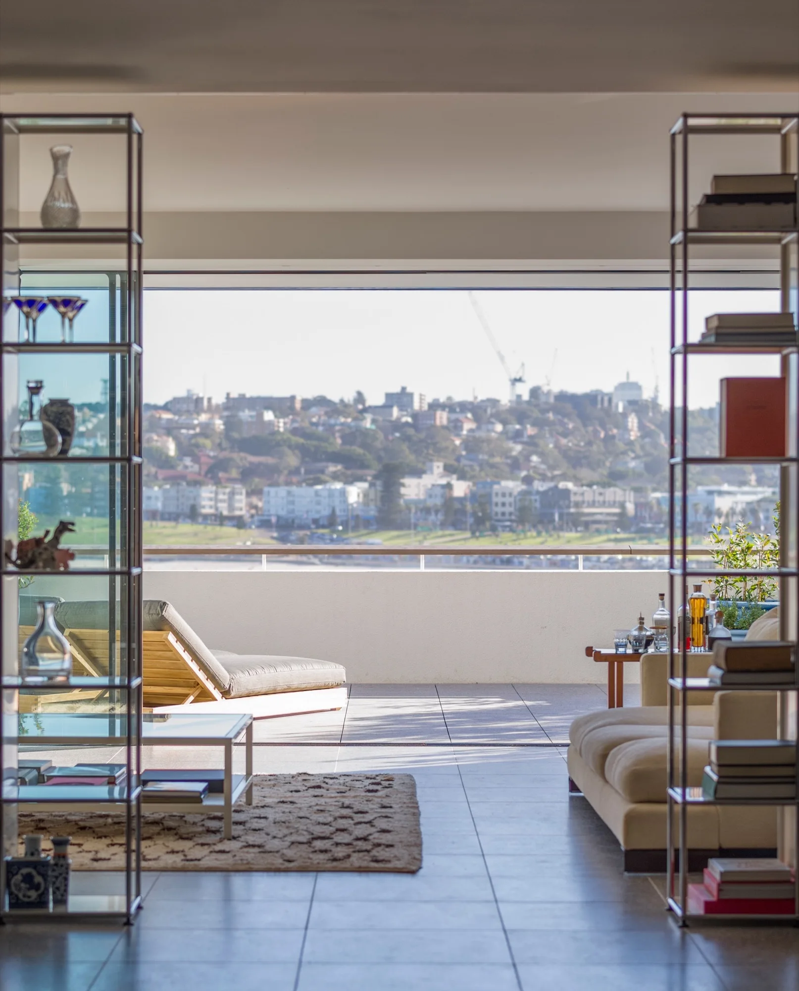 A modern living room features open metal shelving flanking a large sliding glass door that opens to a sunny balcony overlooking a distant city view.