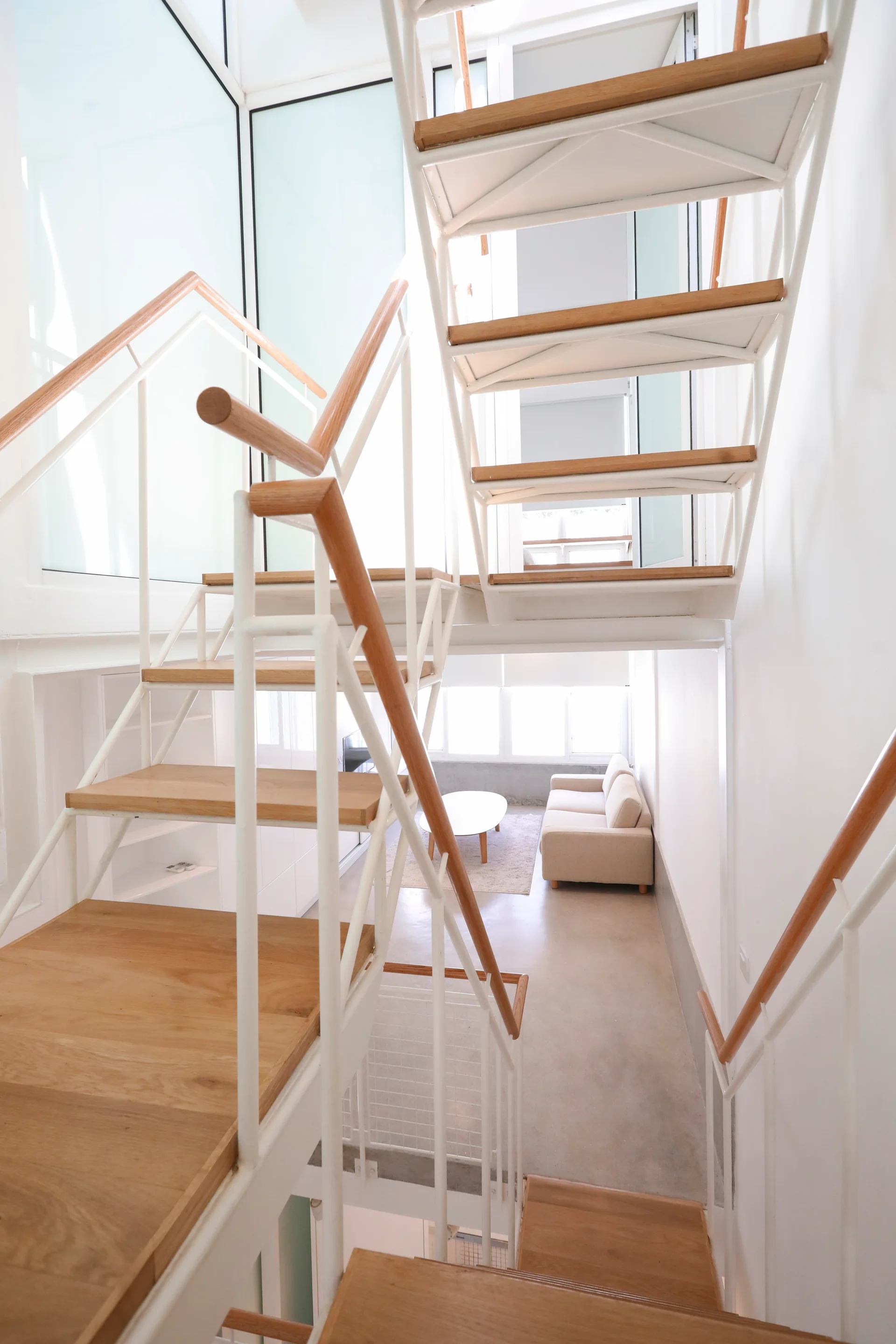 A high-angle view of a modern multi-level staircase with light wood treads and white metal railings overlooking a minimalist living room.