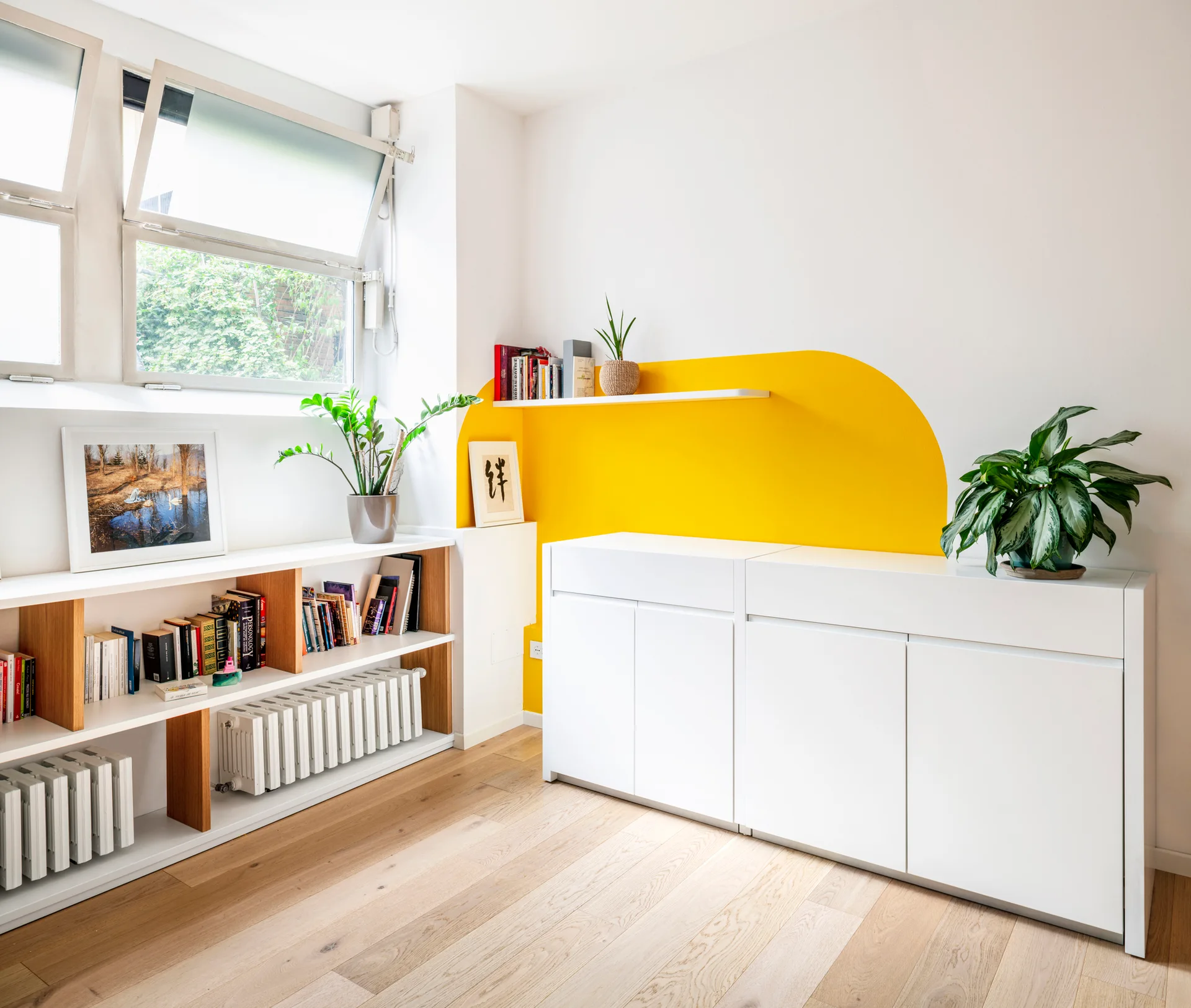 A bright room features white storage cabinets and low shelving under a window, set against a white wall decorated with a yellow painted arch.