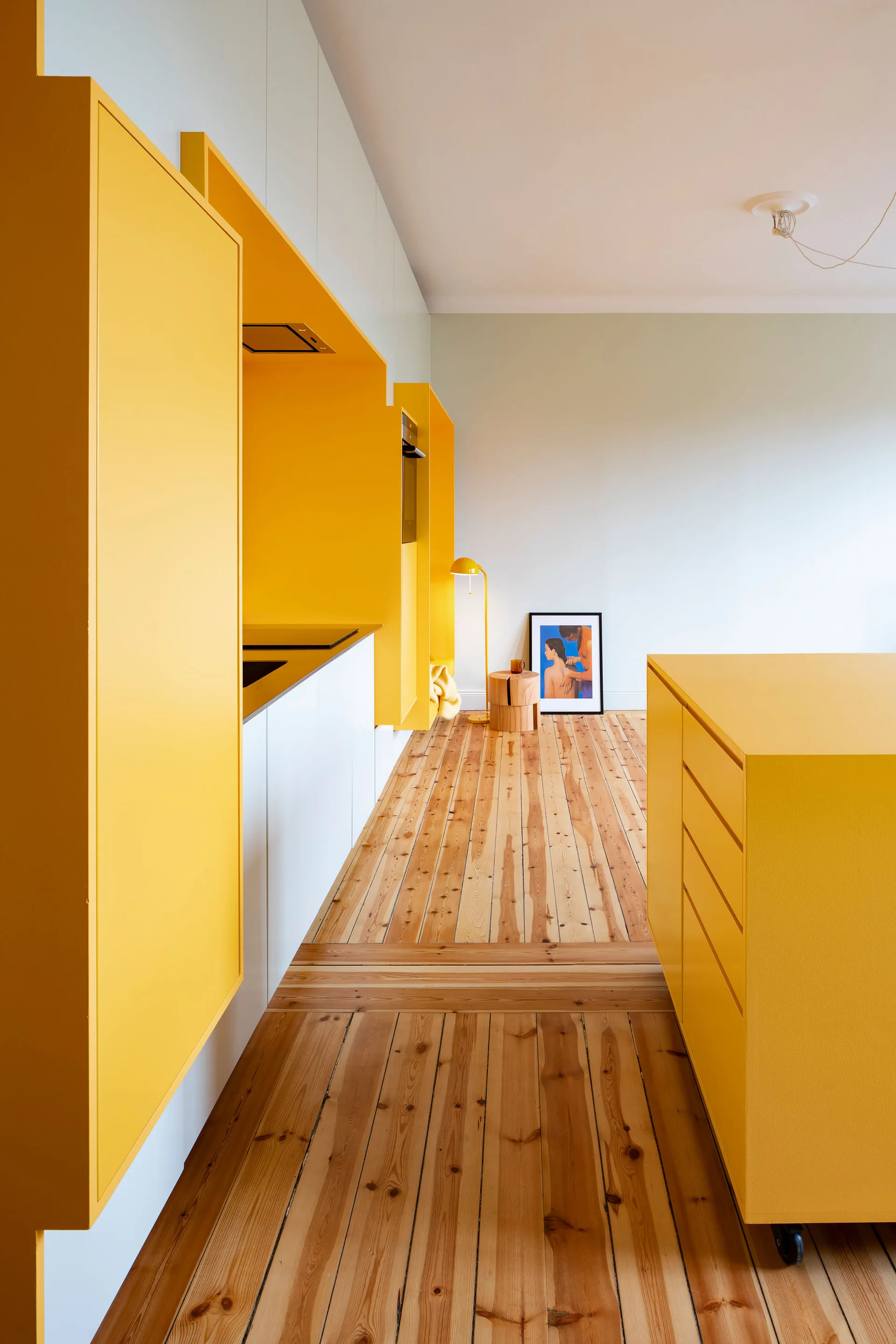A minimalist kitchen features bold matte yellow cabinets, a matching yellow island, and natural wood plank flooring set against white walls.