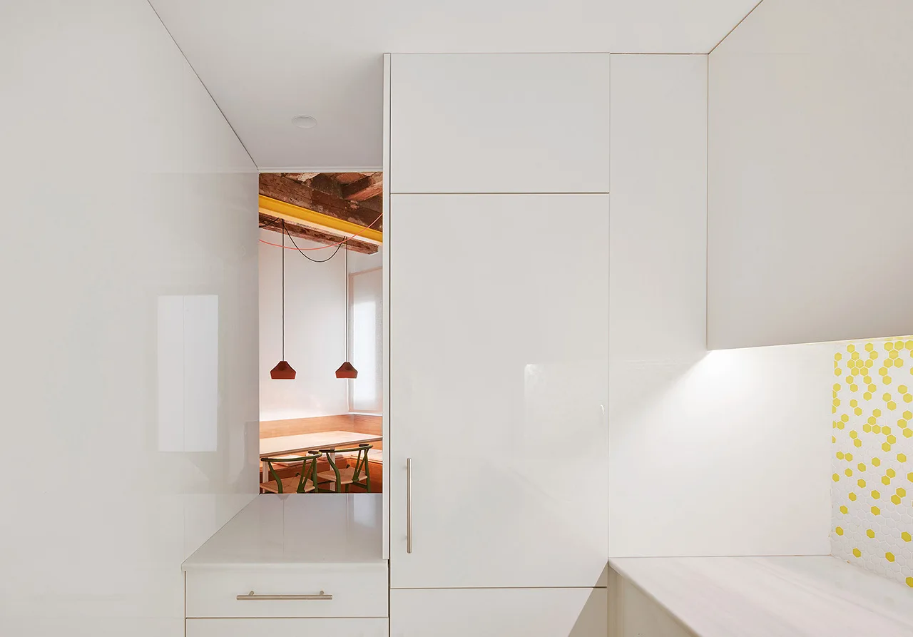 Glossy white kitchen cabinets frame a rectangular opening that looks into a dining room with exposed wood beams and red pendant lights.