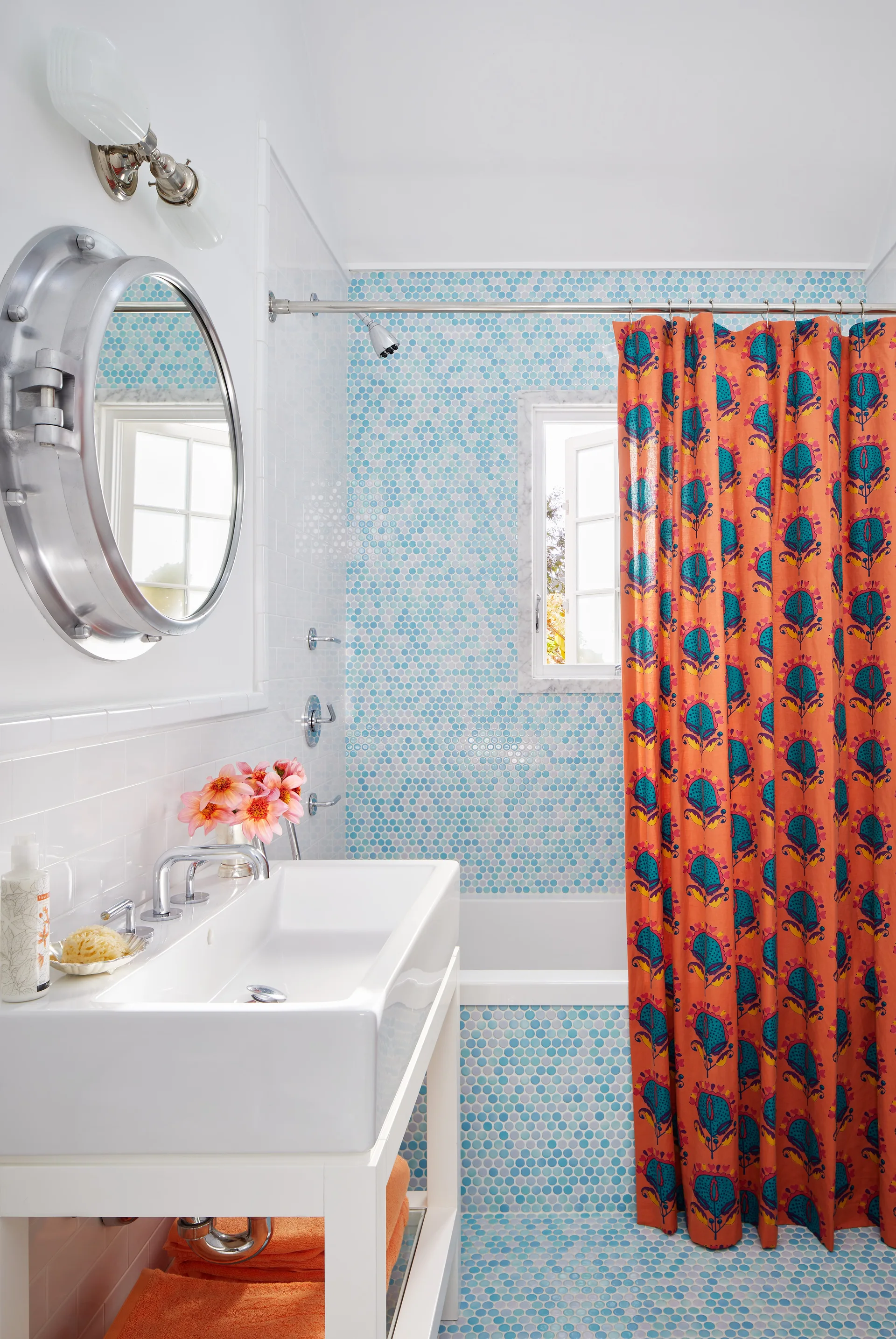 A bathroom features blue penny tile walls and floors, a white console vanity sink, a round porthole mirror, and an orange patterned shower curtain.