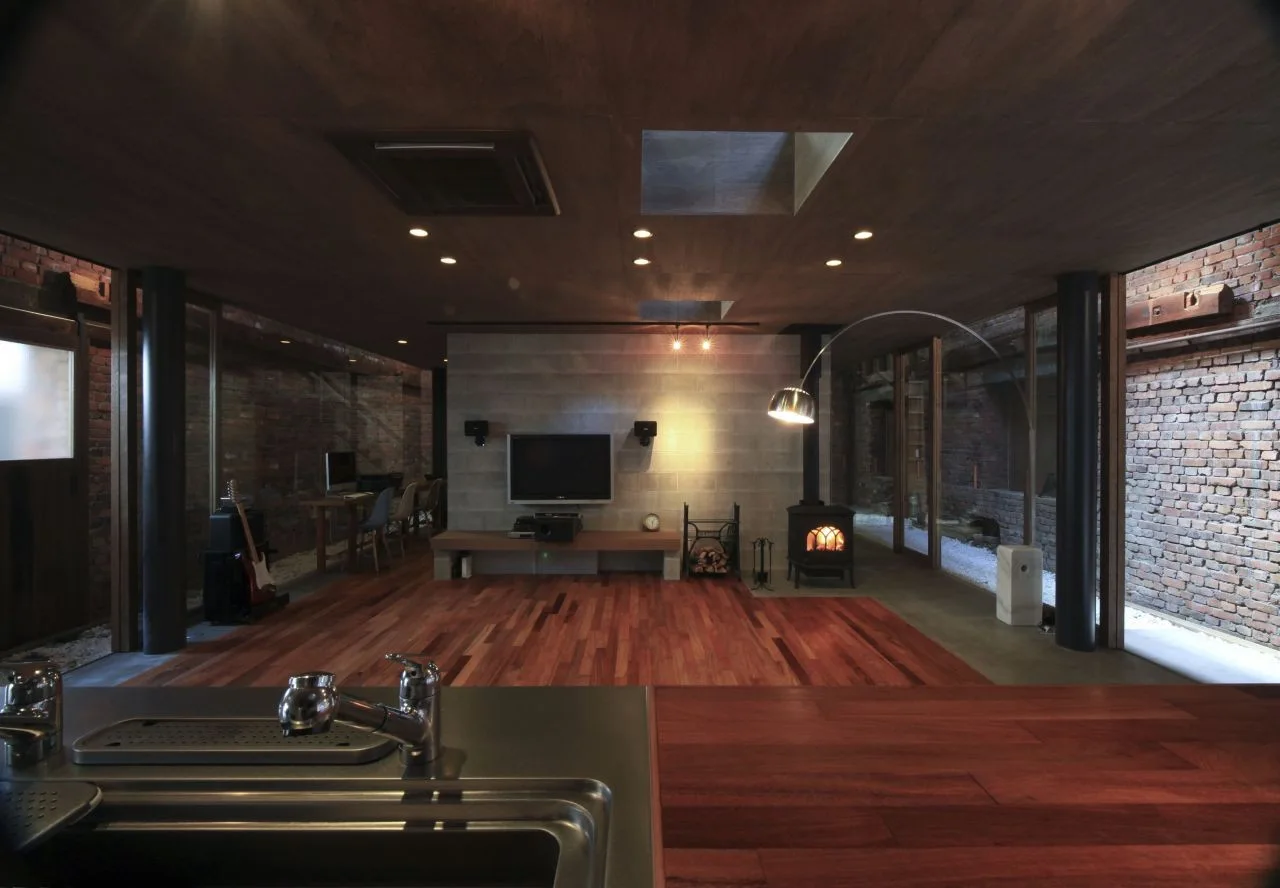 An open-concept living space features a central concrete wall with a television, wood flooring, a dark wood ceiling, and glass walls showing brick.