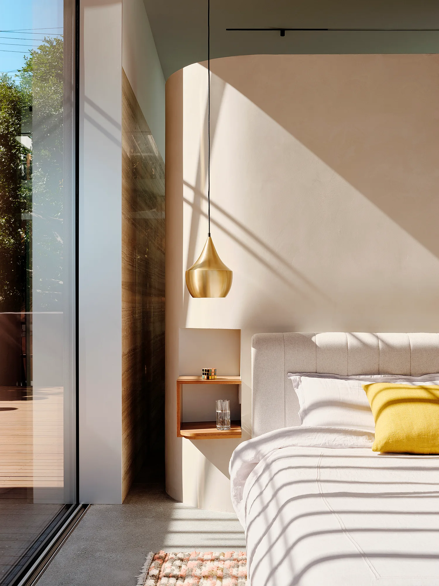 A minimalist bedroom features an upholstered bed, a curved beige plaster wall with a recessed wooden shelf, and a hanging gold pendant light.