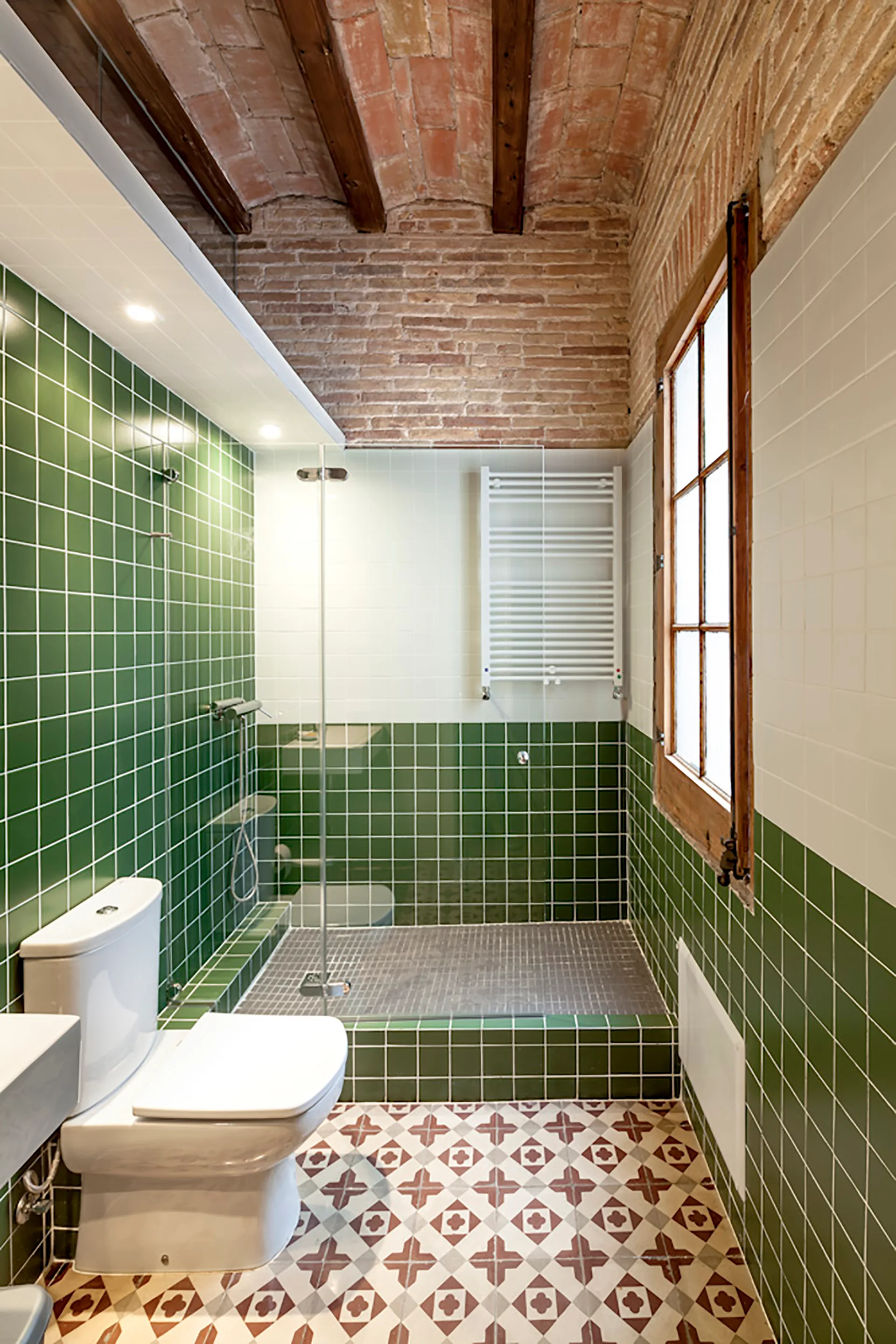 A bathroom featuring green and white tiled walls, a patterned tile floor, a glass shower stall, and an exposed brick vaulted ceiling.