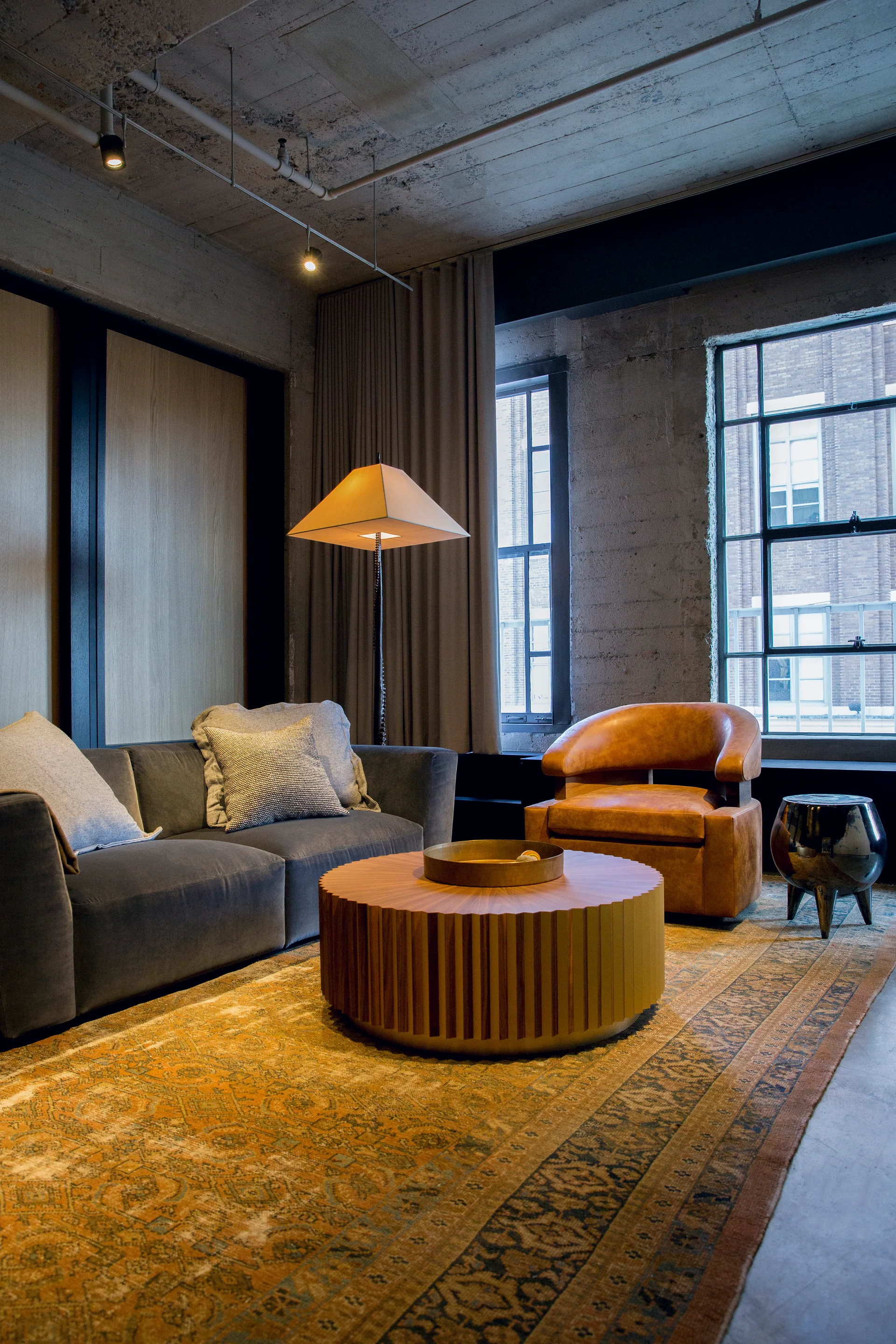 An industrial loft living room with a grey sofa, a fluted round wooden coffee table, a tan leather armchair, and an orange patterned rug.