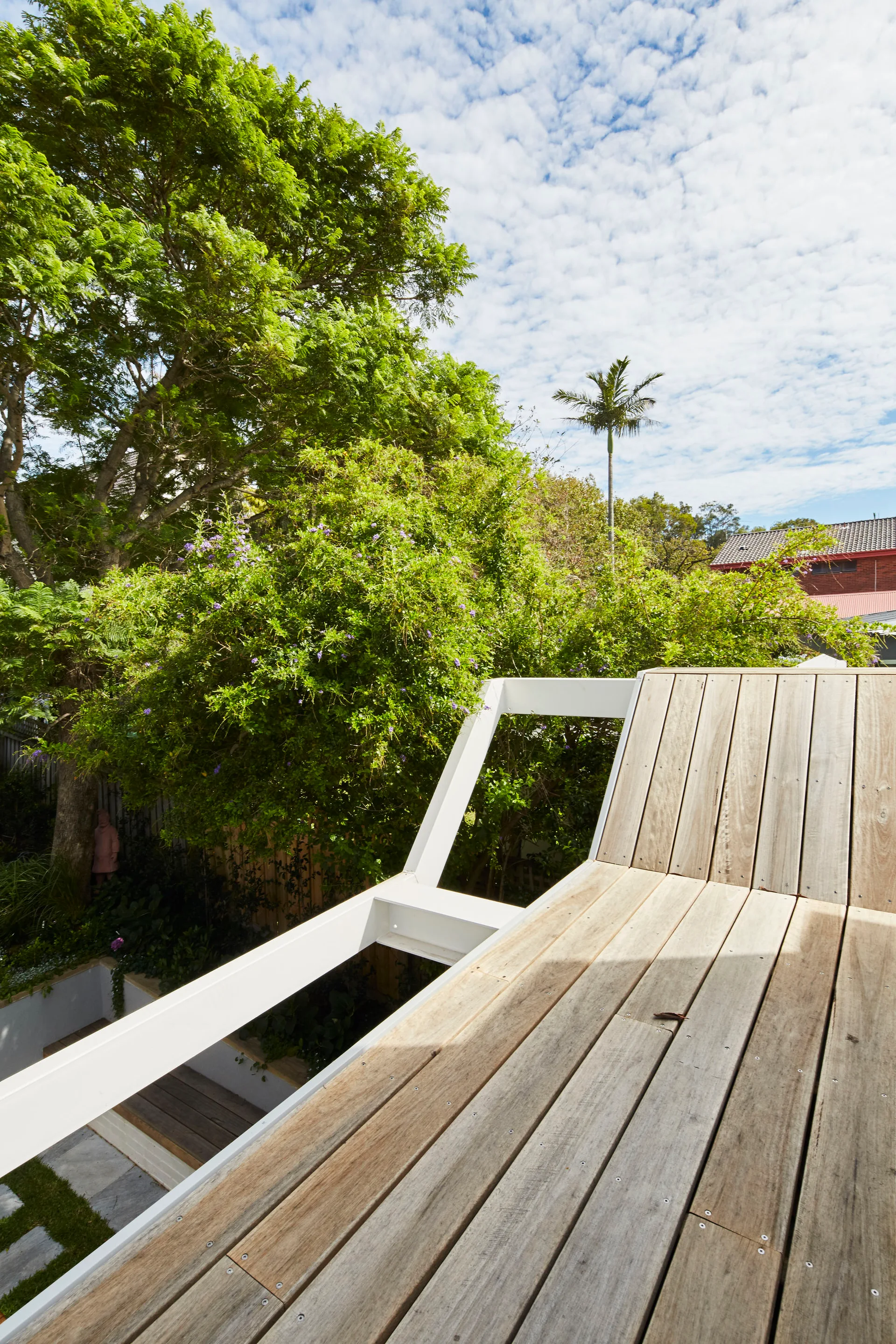 An elevated wood terrace with weathered timber planks and a white metal railing overlooking a lush garden under a blue sky.