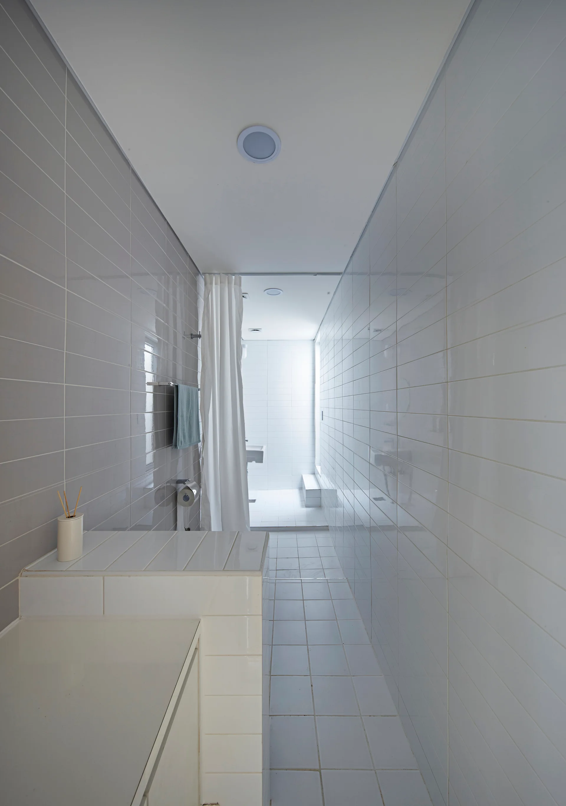 A narrow bathroom with glossy grey tiles on one wall, white tiles on the opposite wall, white floor tiles, and a white shower curtain.