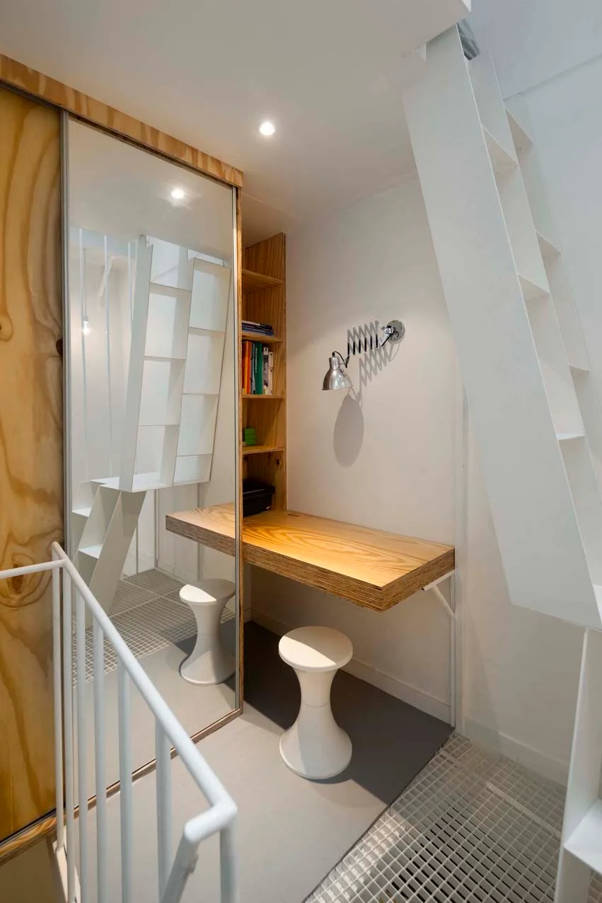 A compact home office features a built-in plywood desk, a matching wooden bookshelf, a white hourglass stool, and a tall sliding mirror next to a staircase.