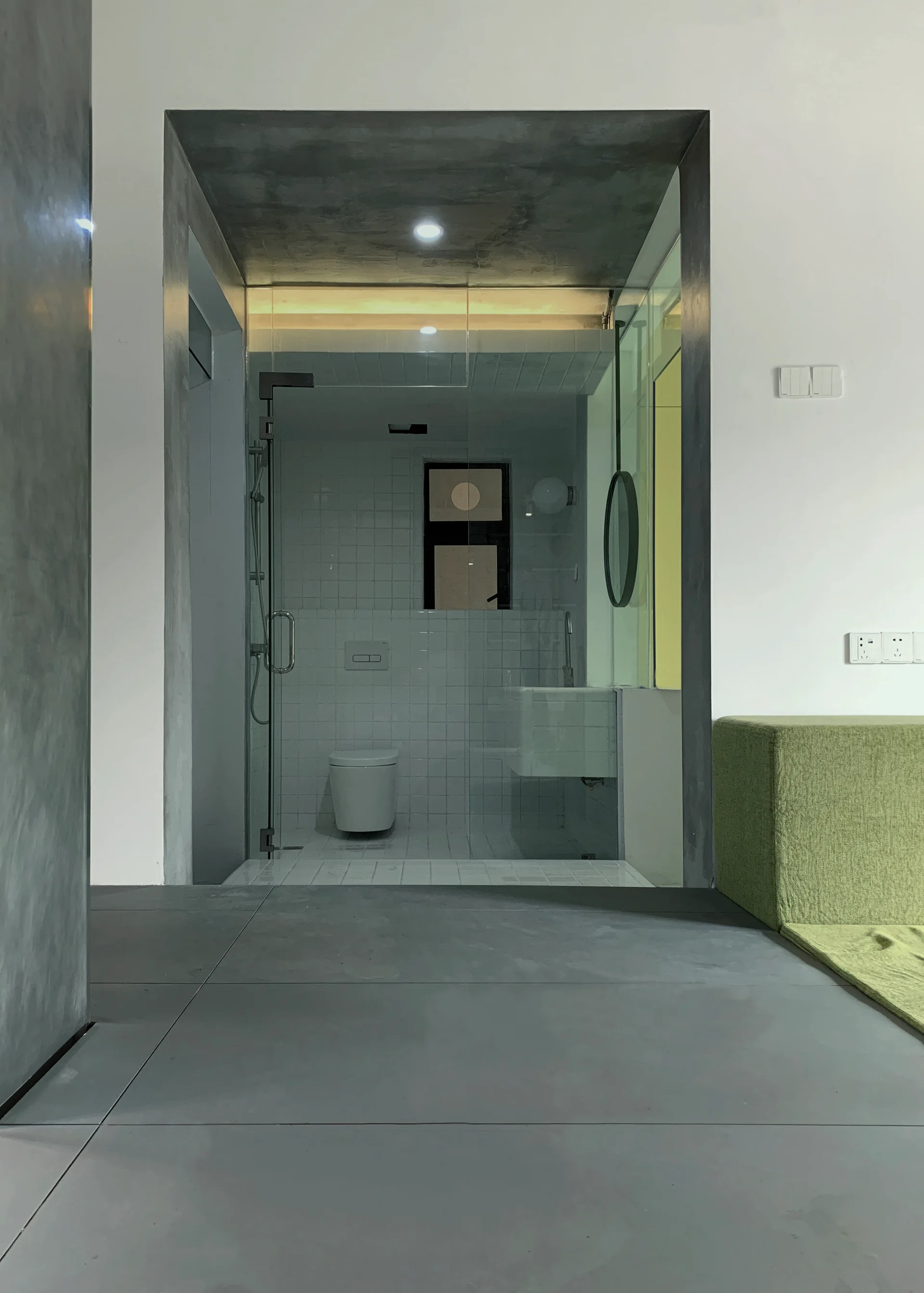 A modern bathroom with white grid tiles and a toilet is visible through a glass partition and framed by a dark gray concrete entryway next to a green sofa.