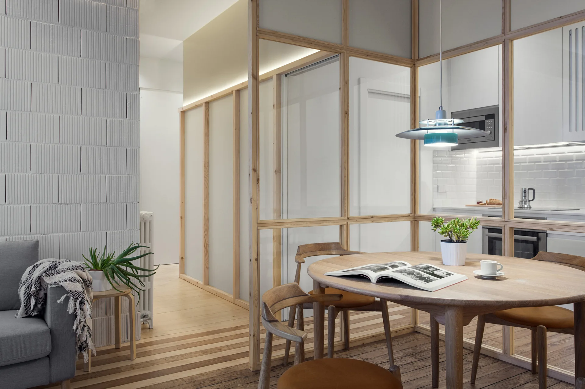 A round wooden dining table and chairs sit next to a light wood-framed glass partition separating the space from a white kitchen.