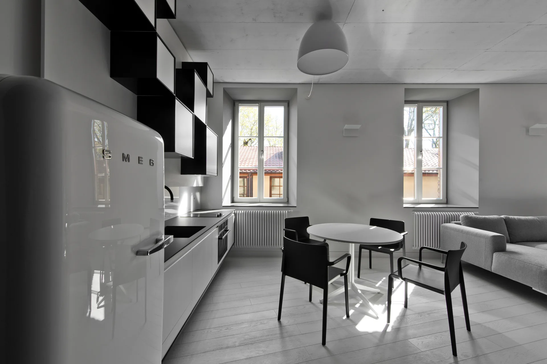 A minimalist black and white kitchen featuring a retro white refrigerator, modular wall shelves, a round dining table with chairs, and two windows.