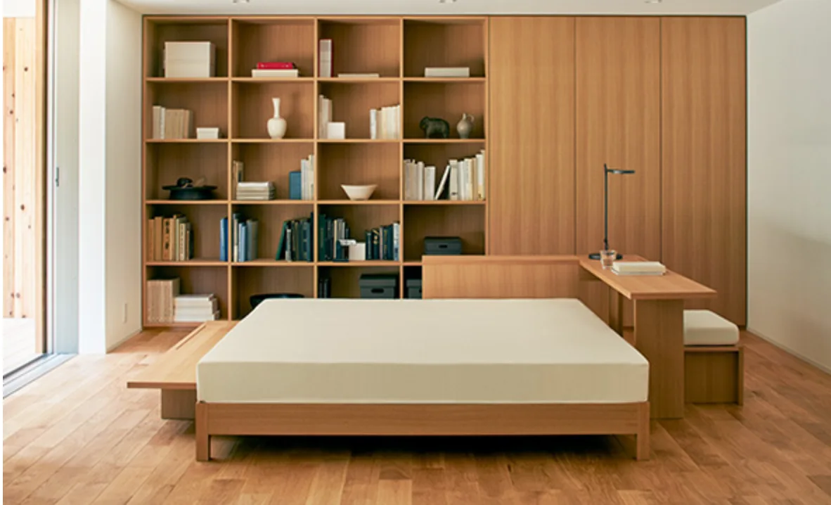 A minimalist bedroom featuring a low wood platform bed integrated with a built-in light wood desk and a wall-to-wall grid bookcase.