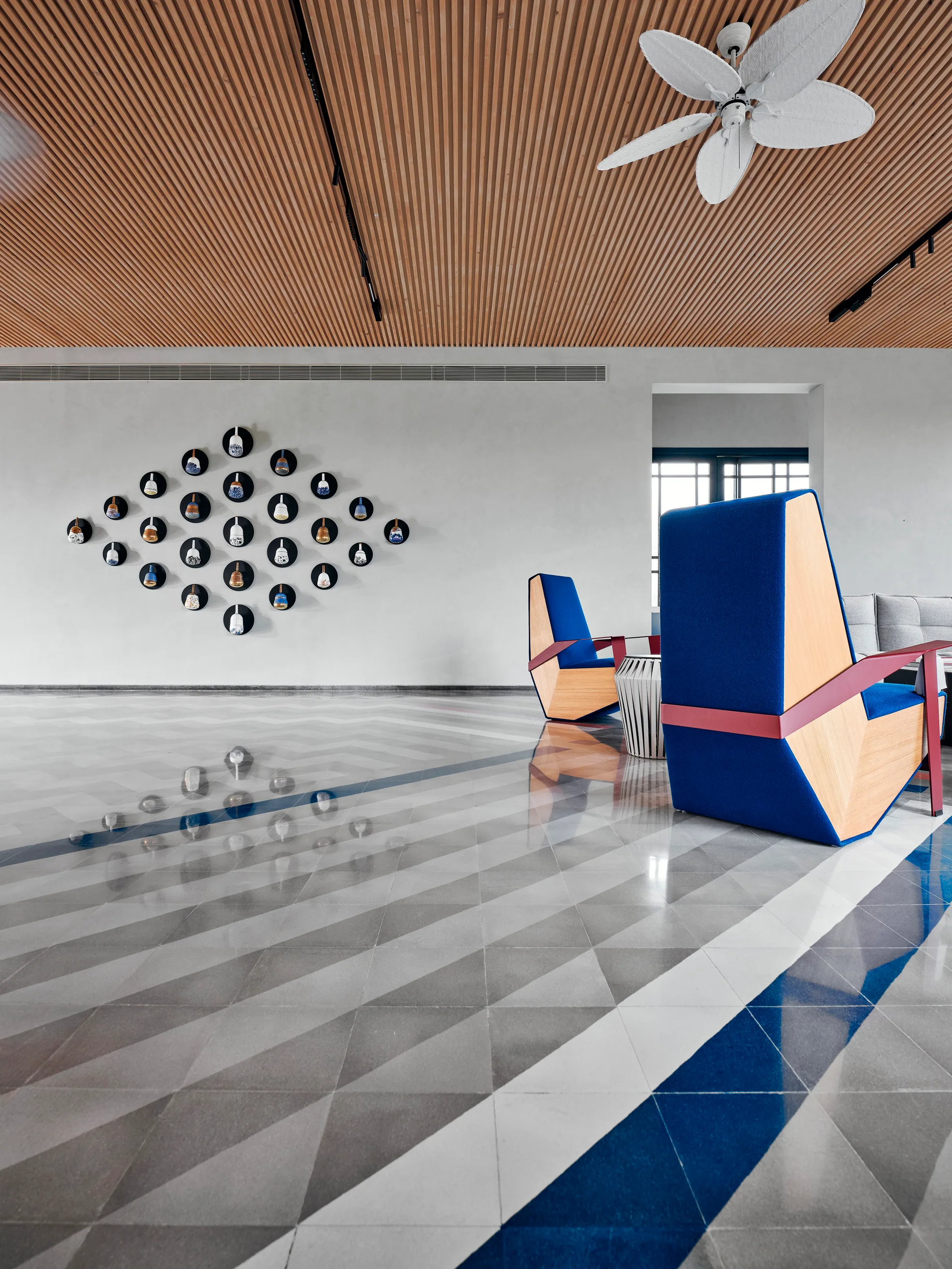 A modern lounge features blue geometric armchairs on polished triangular tile floors beneath a slatted wood ceiling and a diamond-shaped wall display.
