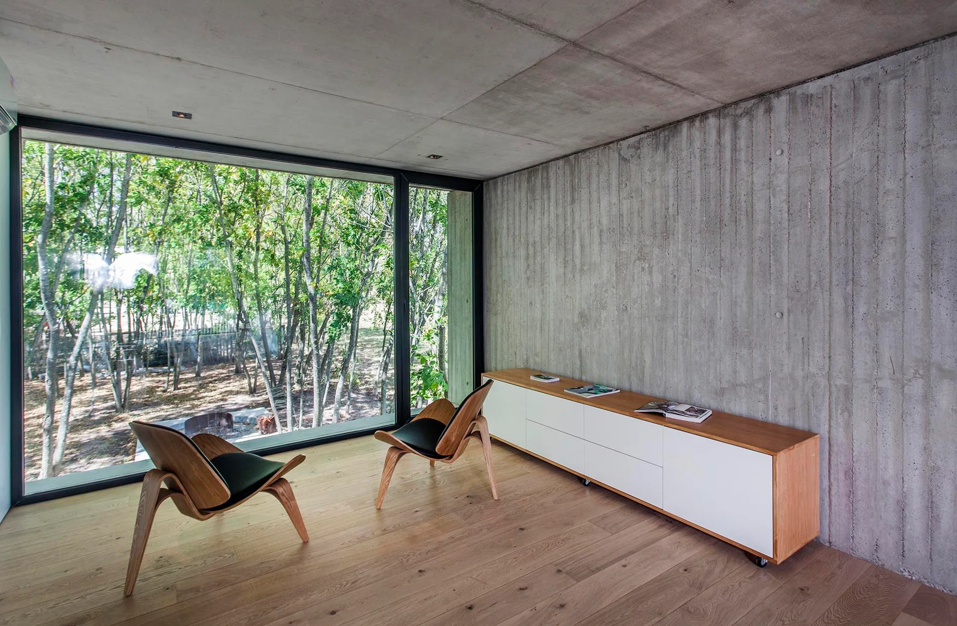 Two curved wooden lounge chairs sit on a light wood floor near a long white console and an exposed concrete wall with a large forest-view window.